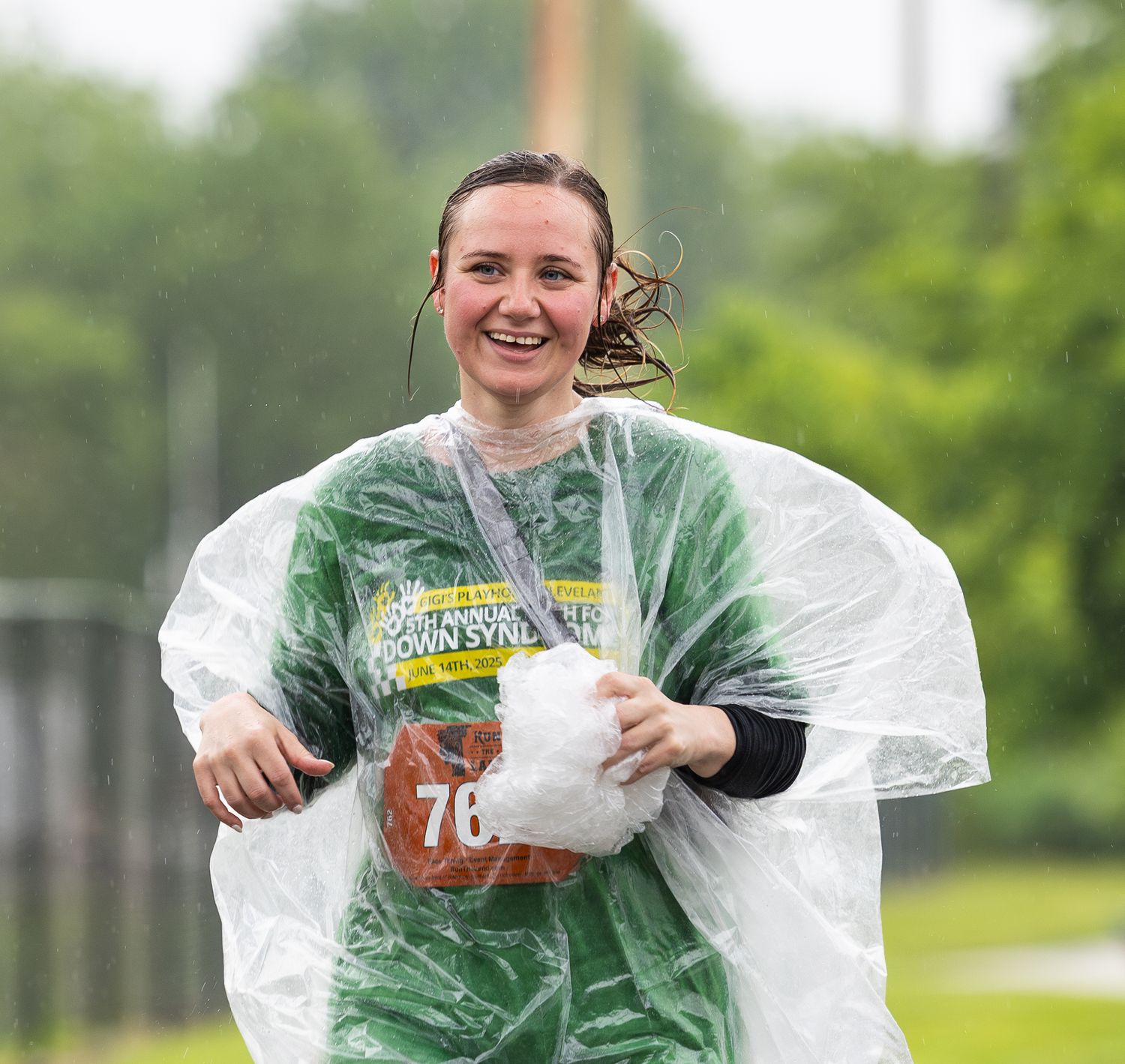 Person in green rain poncho smiling and holding groceries while walking in the rain.
