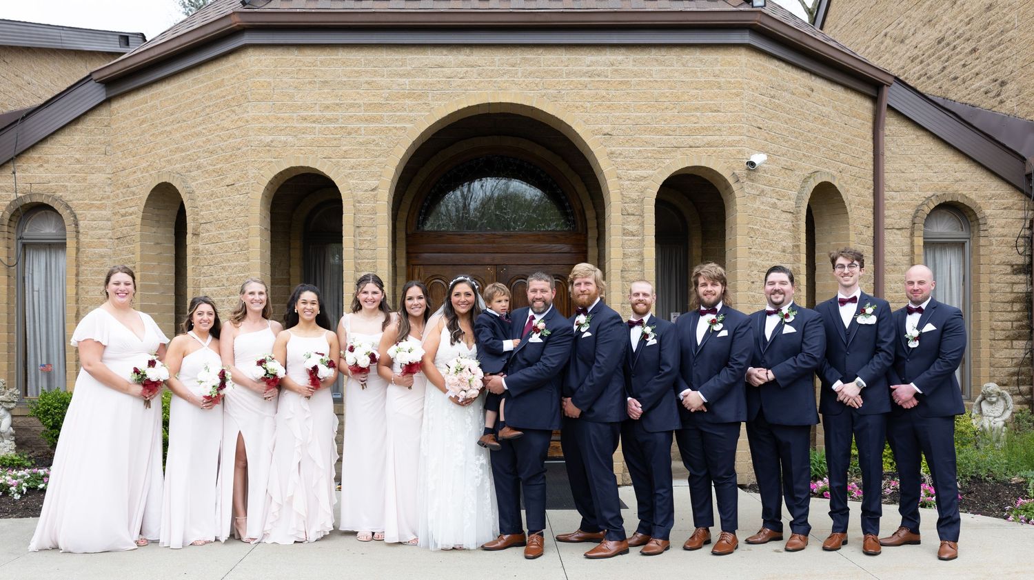 Wedding party members in white dresses and navy suits pose in front of a stone building for formal photos.