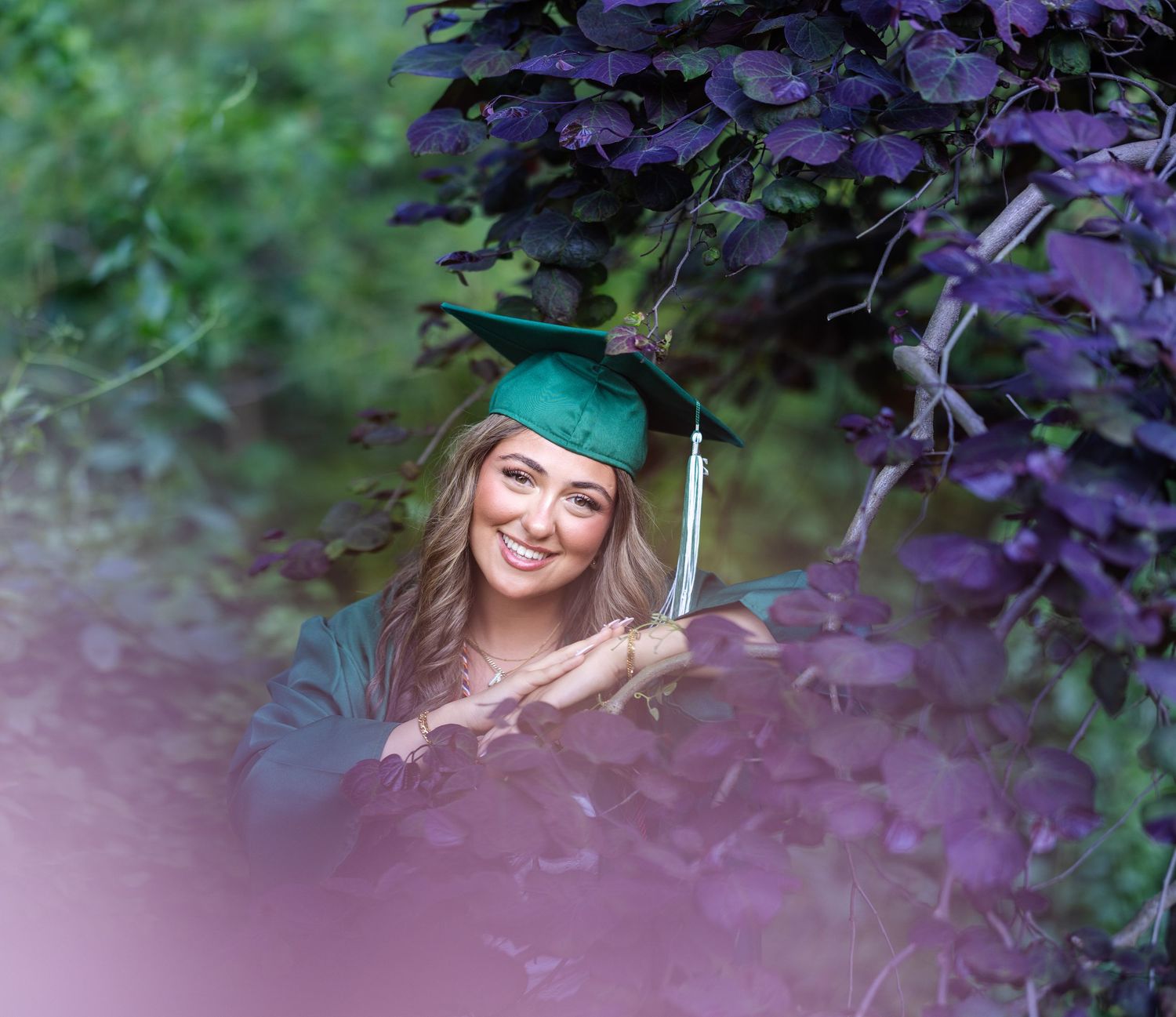 Smiling graduate in green cap and gown posing among purple foliage for senior graduation photos.