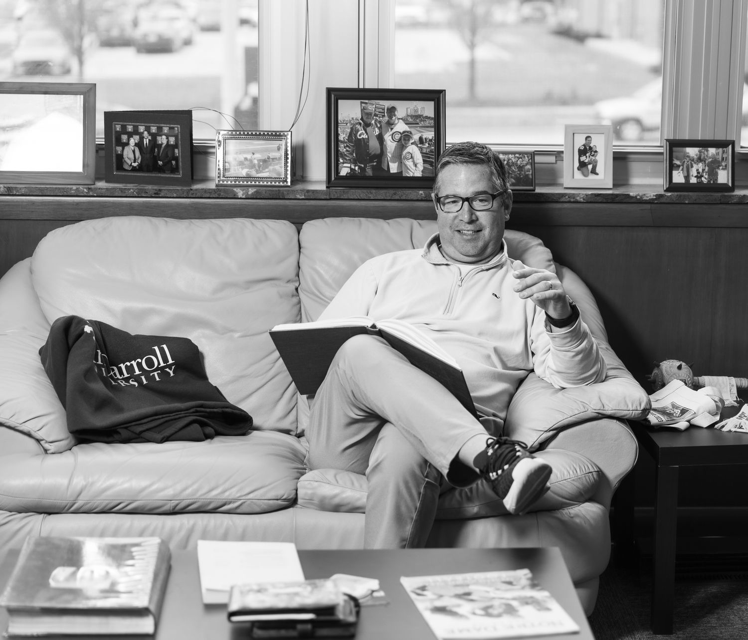 Black and white photo of someone relaxing on a couch with a drink, surrounded by framed photos on a shelf behind them.