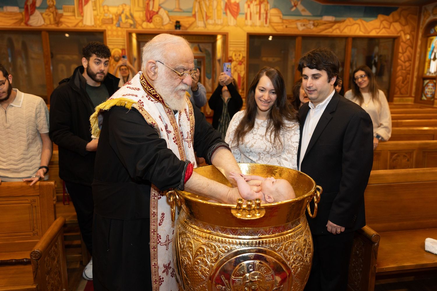 Orthodox Christian baptism ceremony in a church with priest and family gathered around ornate gold baptismal font.
