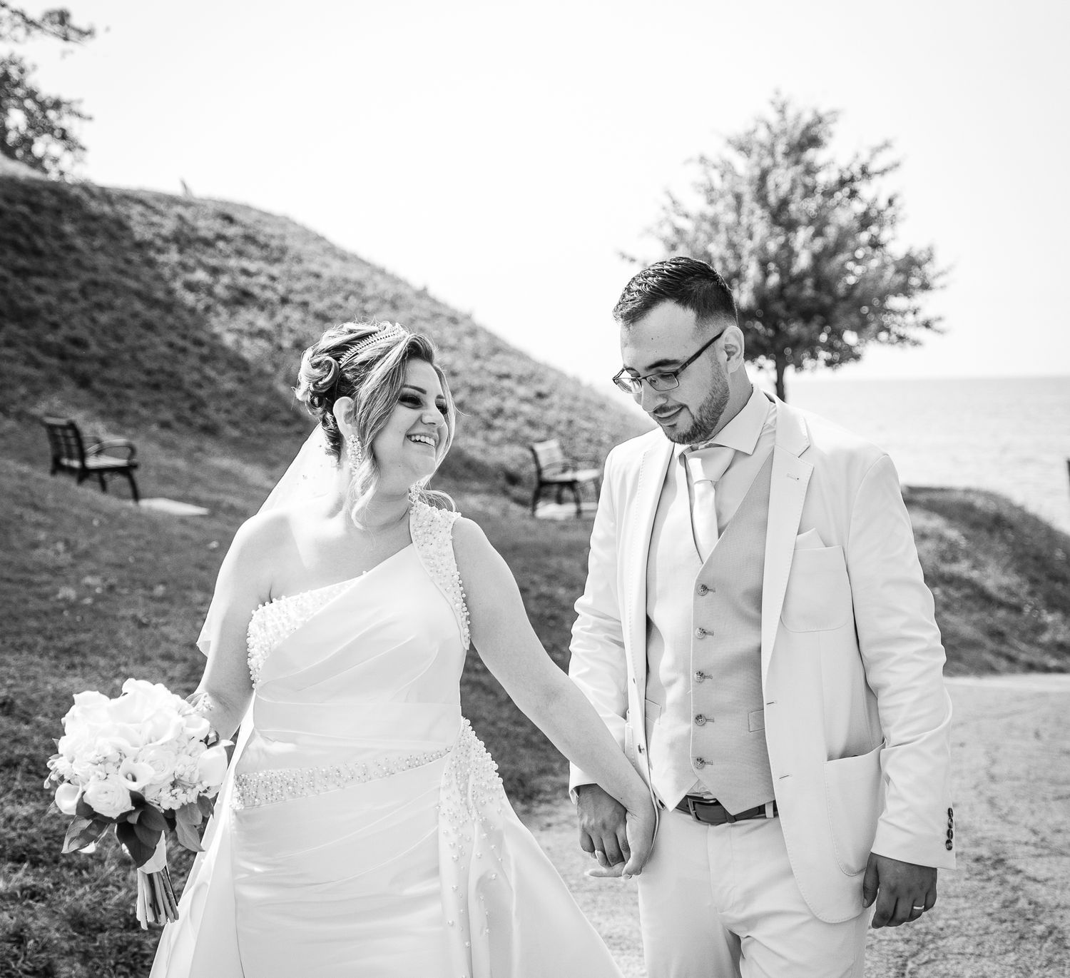 A couple in white wedding attire walks together on a beach path while holding hands and laughing.