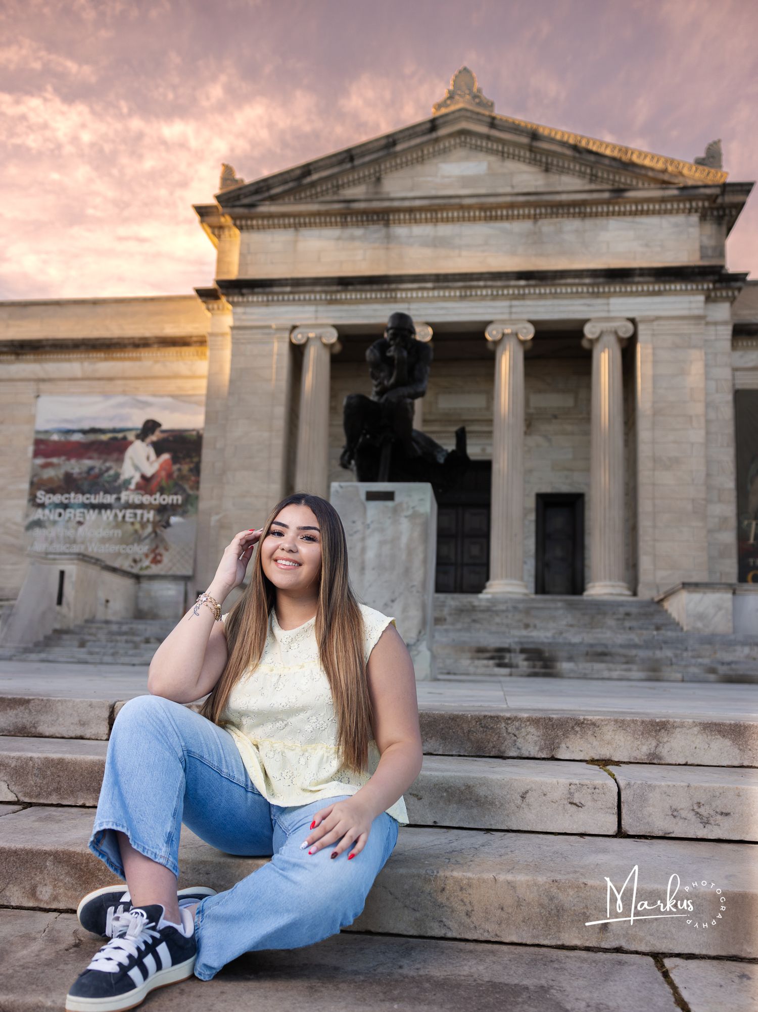 Young woman sitting on steps before a neoclassical building at sunset, smiling casually in jeans and a yellow top.