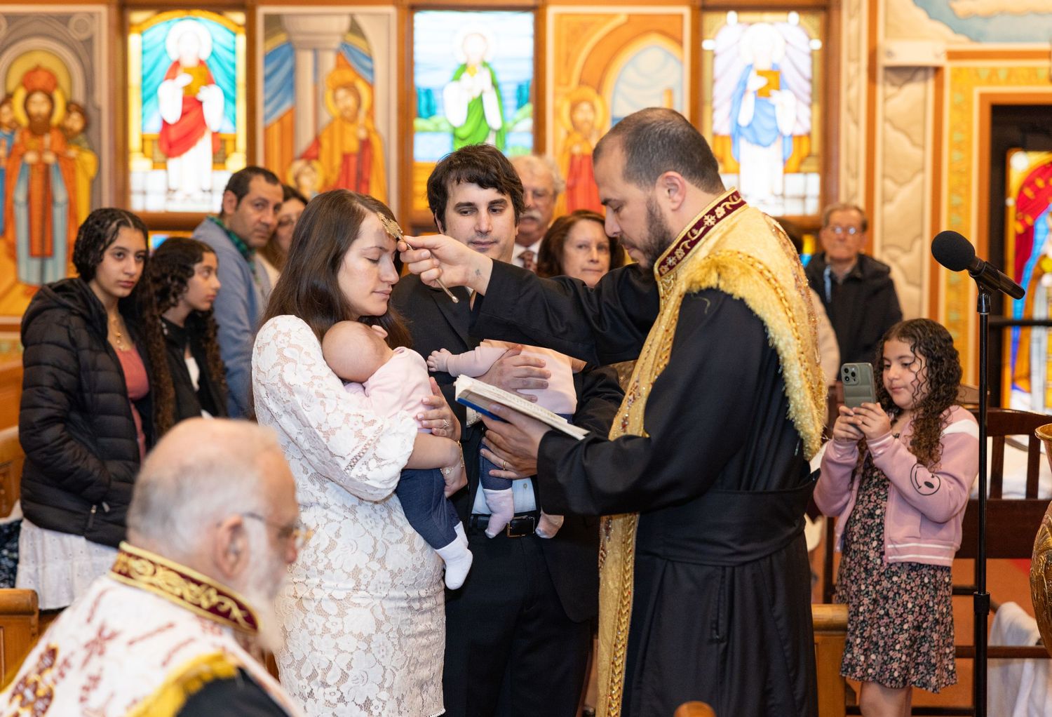 A religious baptism ceremony takes place inside an Orthodox church with stained glass windows and icons in the background.