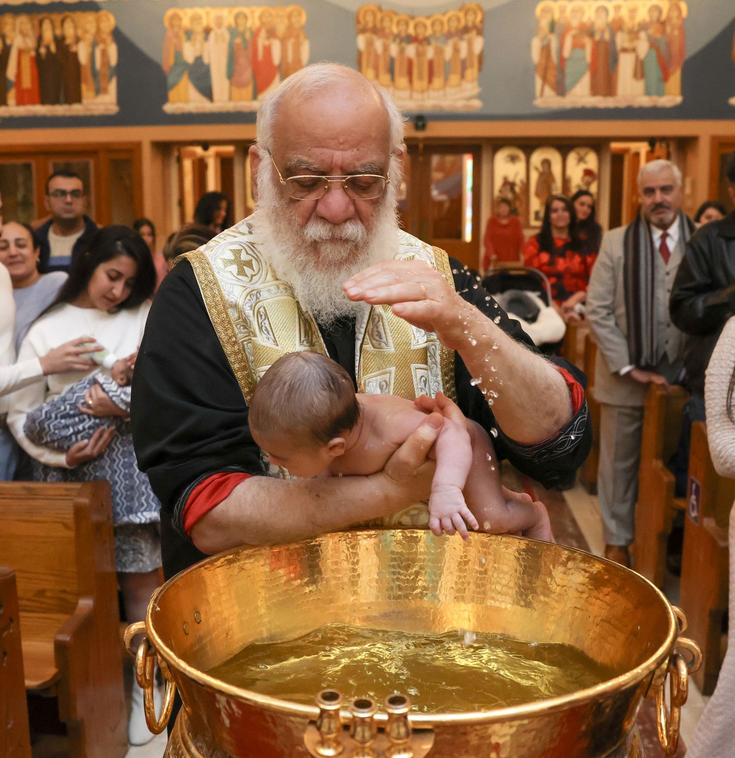 Orthodox priest in black robes performs baptism ceremony with large golden baptismal font in ornate church setting.