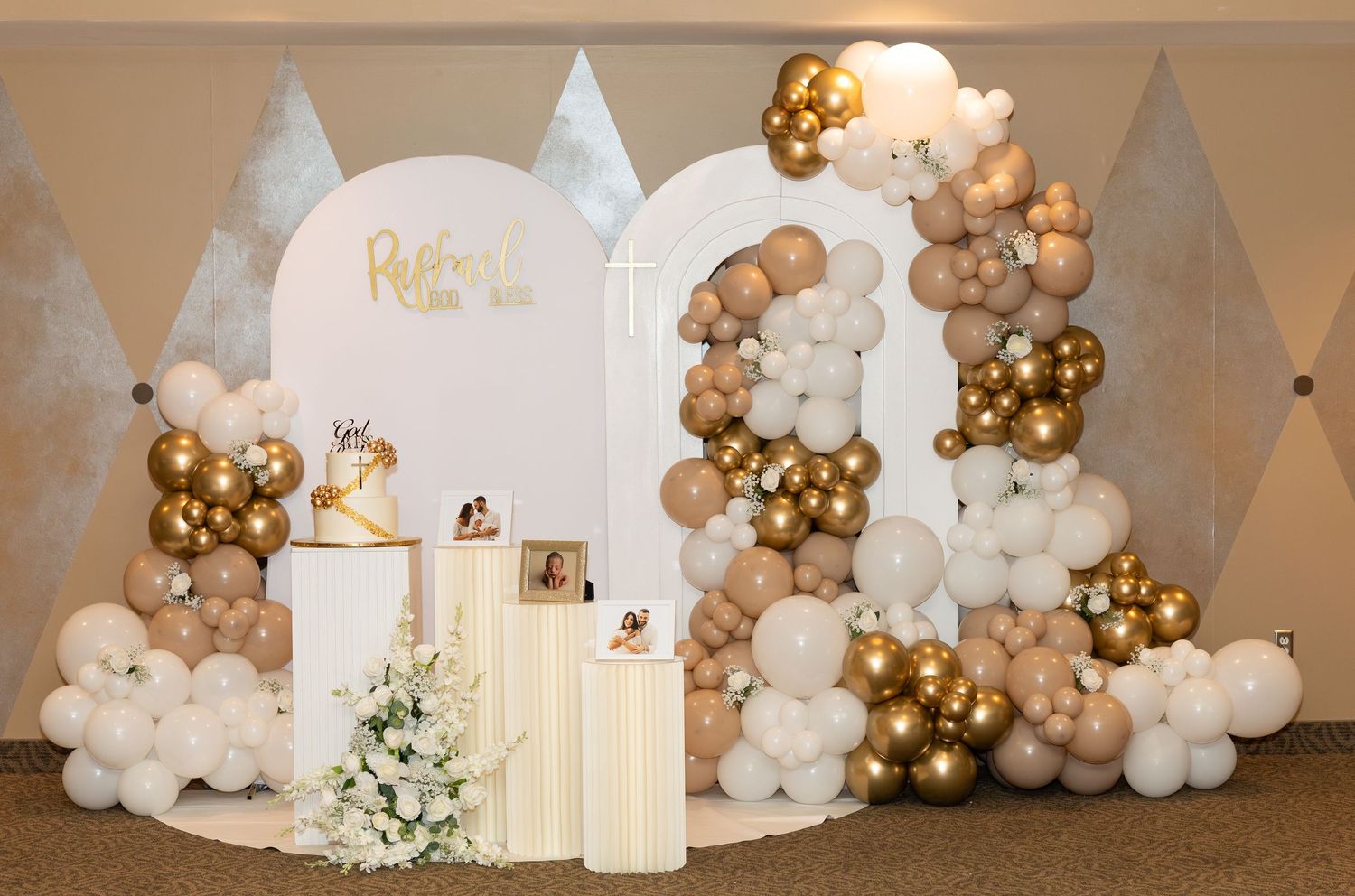 Elegant wedding backdrop with white and gold balloon arch garland and decorative candle pillars against a beige wall.
