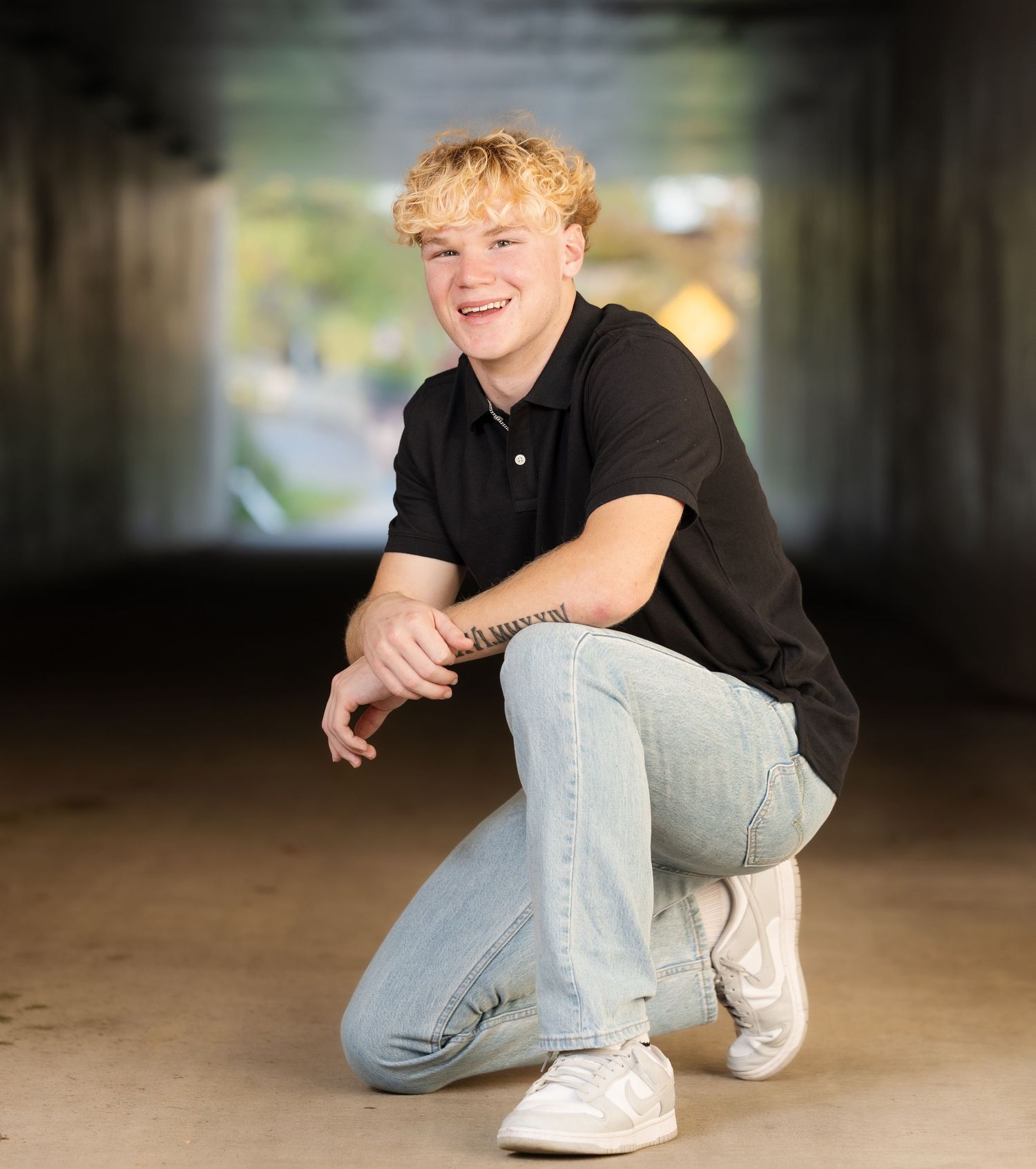 Person in black shirt and light jeans crouches in a shadowy tunnel-like setting with dramatic lighting.