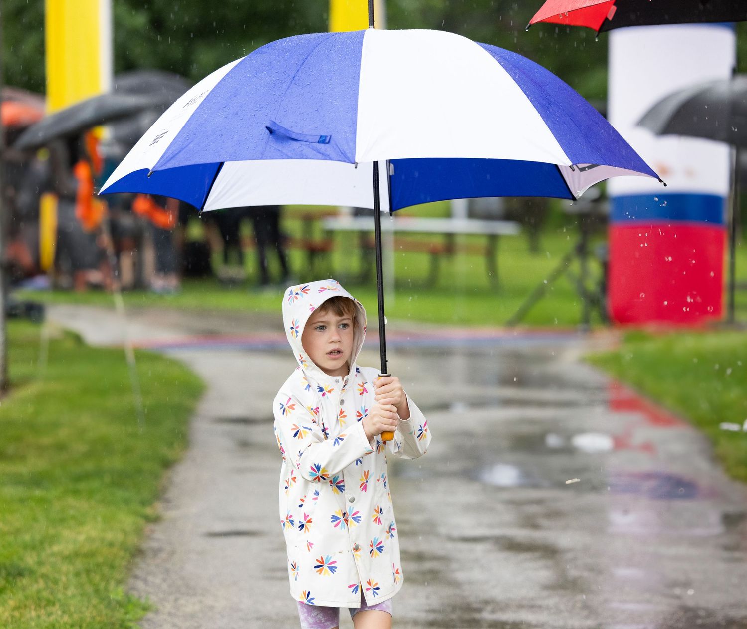 A small figure in a white patterned raincoat holds a blue and white umbrella while standing on a wet path on a rainy day.