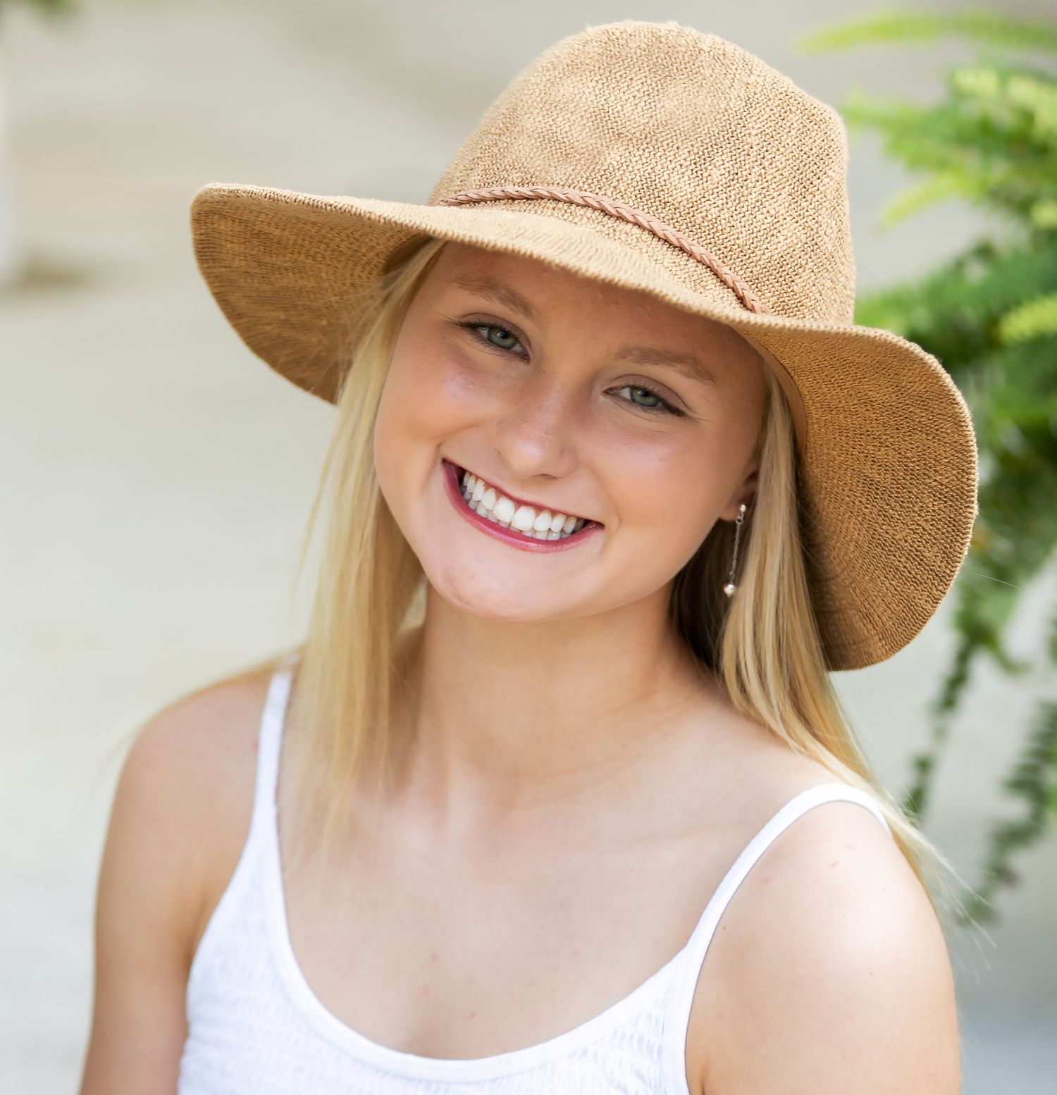 A person wearing a wide-brimmed straw hat and white top smiles outdoors on a sunny day.