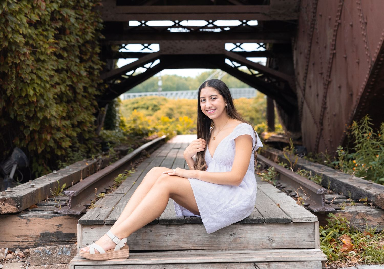 A person in a white dress sits on rustic train tracks under a bridge with autumn foliage in the background.