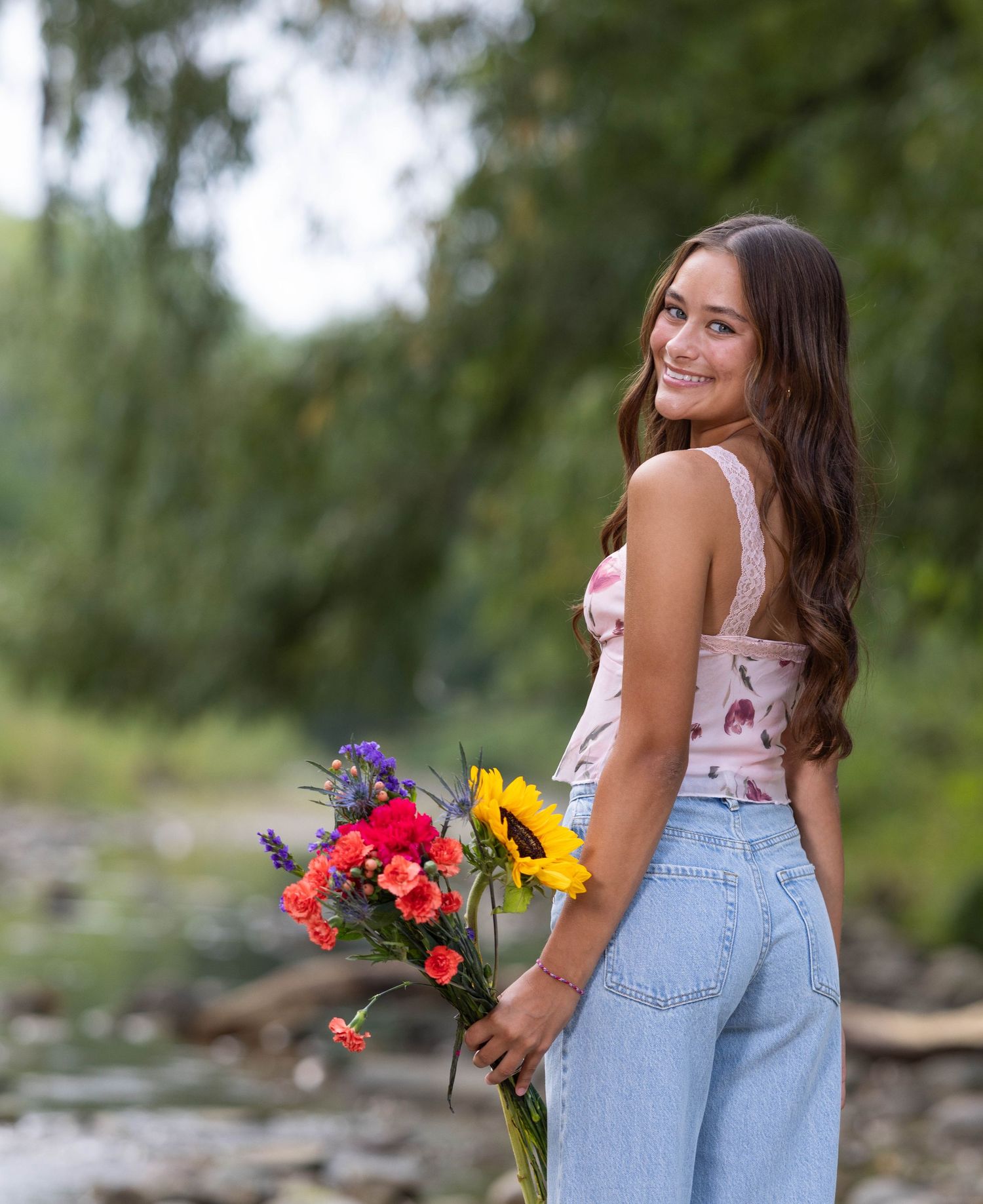 A person in a floral top and jeans holds a vibrant bouquet of wildflowers while posing outdoors in a natural setting.