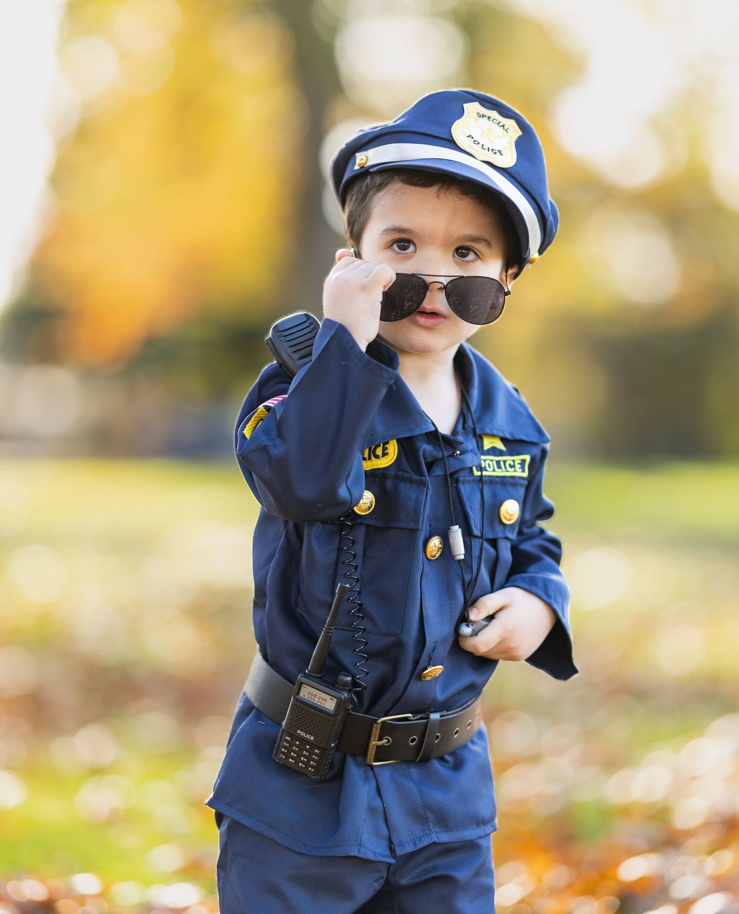 A small child dressed in a police officer costume poses playfully in autumn leaves.