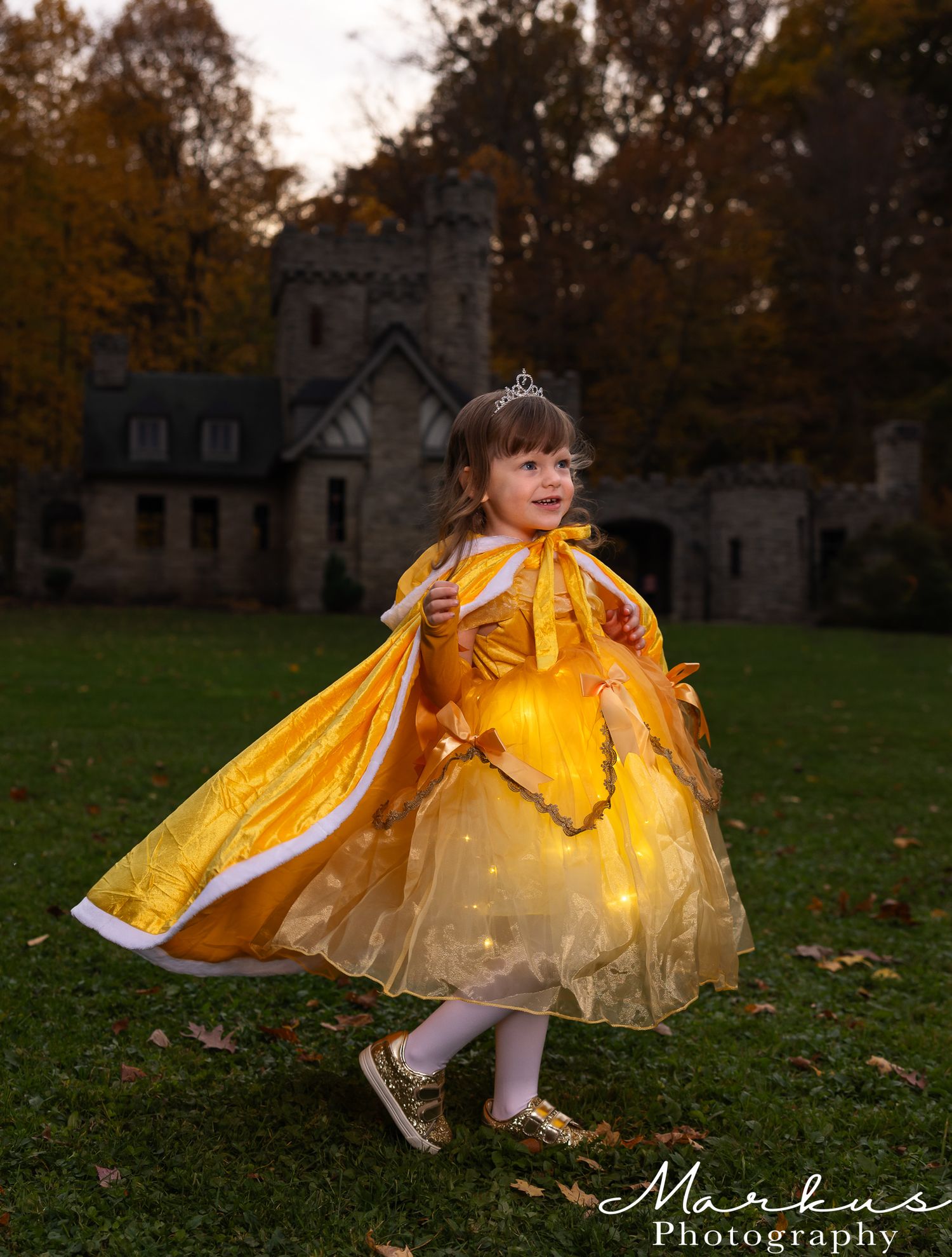 A child in a flowing yellow princess dress twirls and plays in fallen autumn leaves near a castle-like structure.