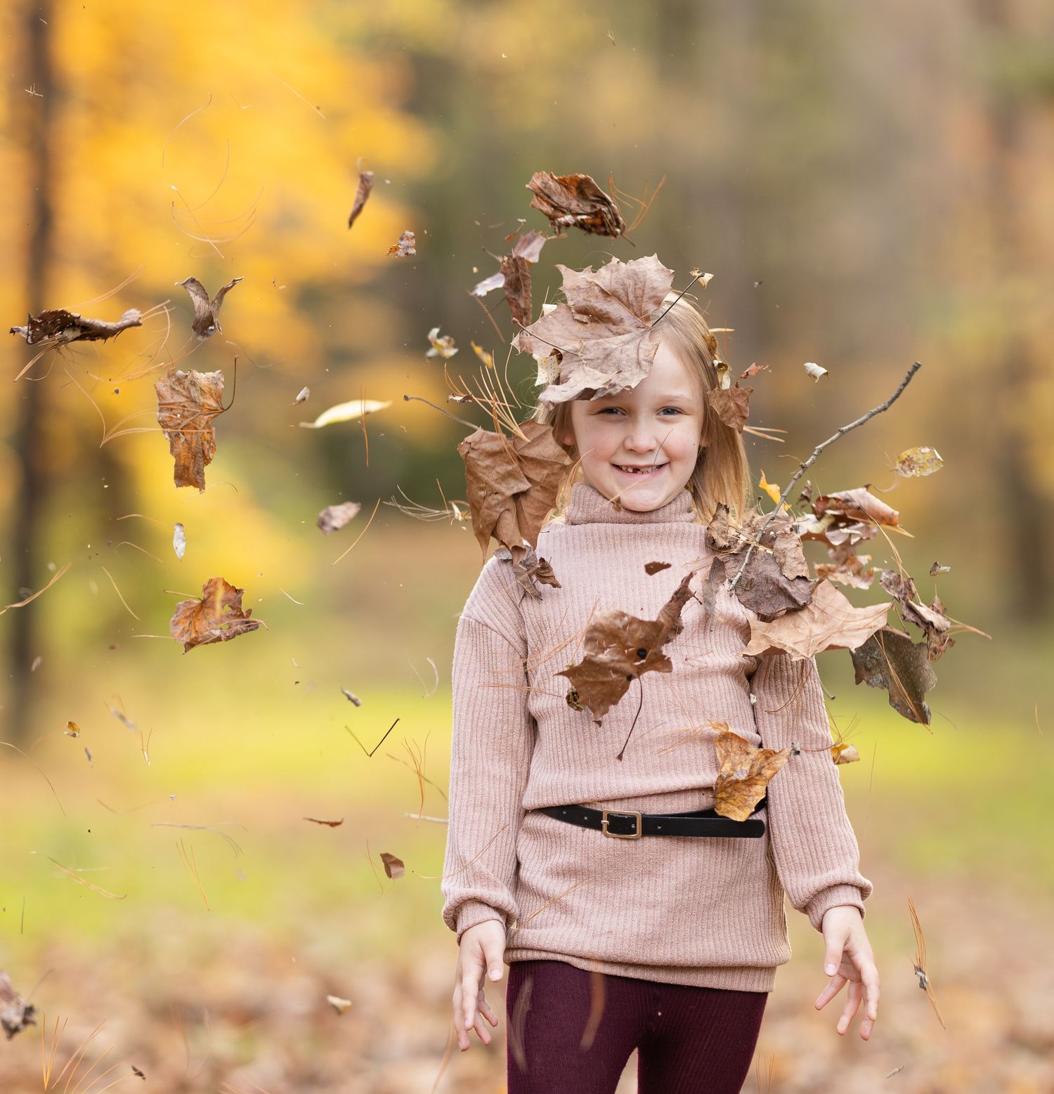 A person in a beige sweater joyfully plays with falling autumn leaves on a sunny fall day.