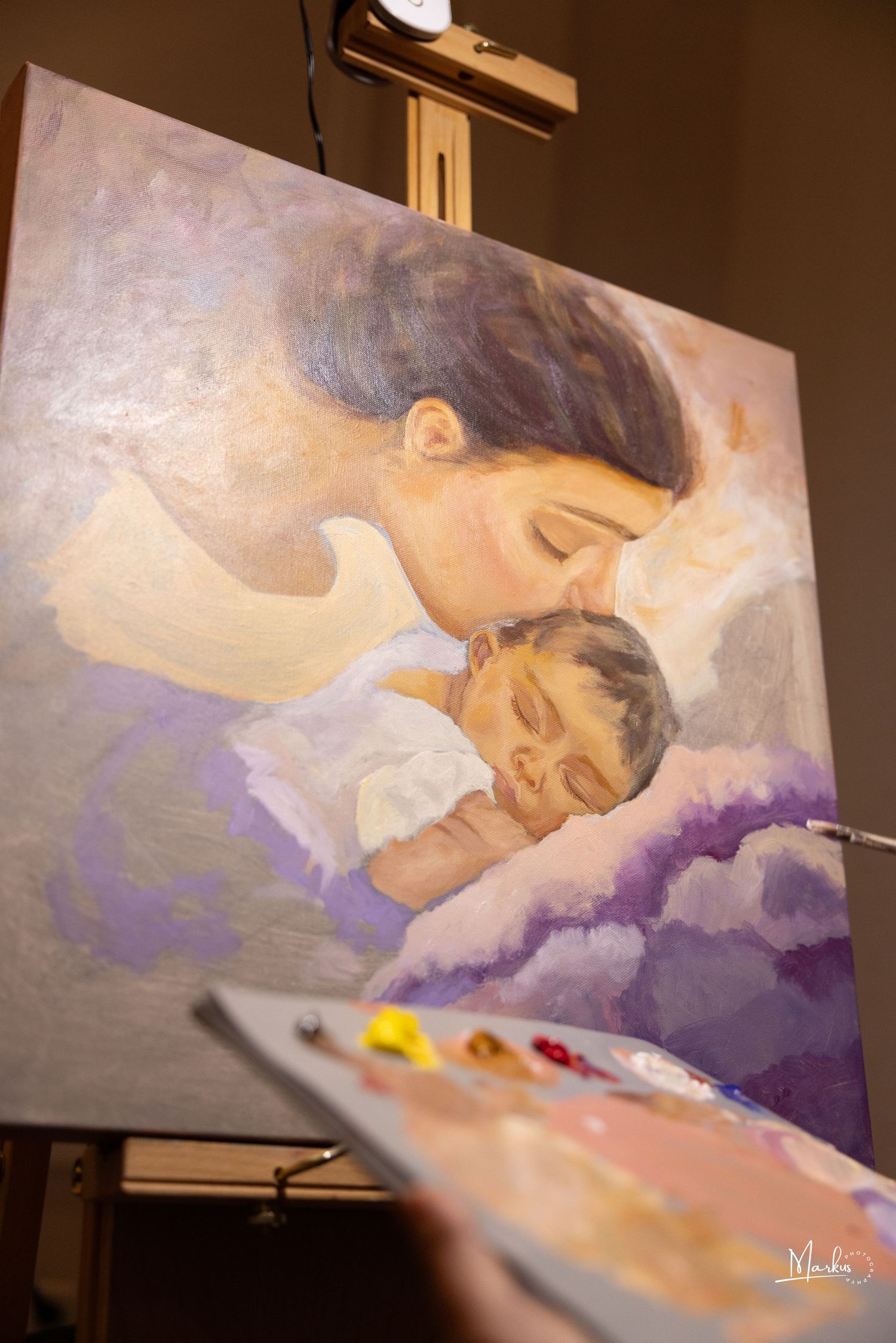 A painting on an easel shows a tender moment between parent and sleeping child in soft purple and beige tones.