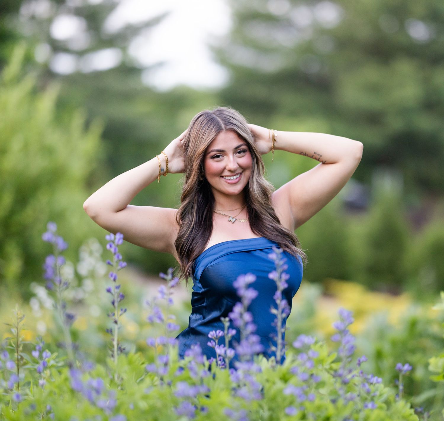 Young woman in blue dress posing among purple wildflowers in a lush garden, smiling with hands behind her head.