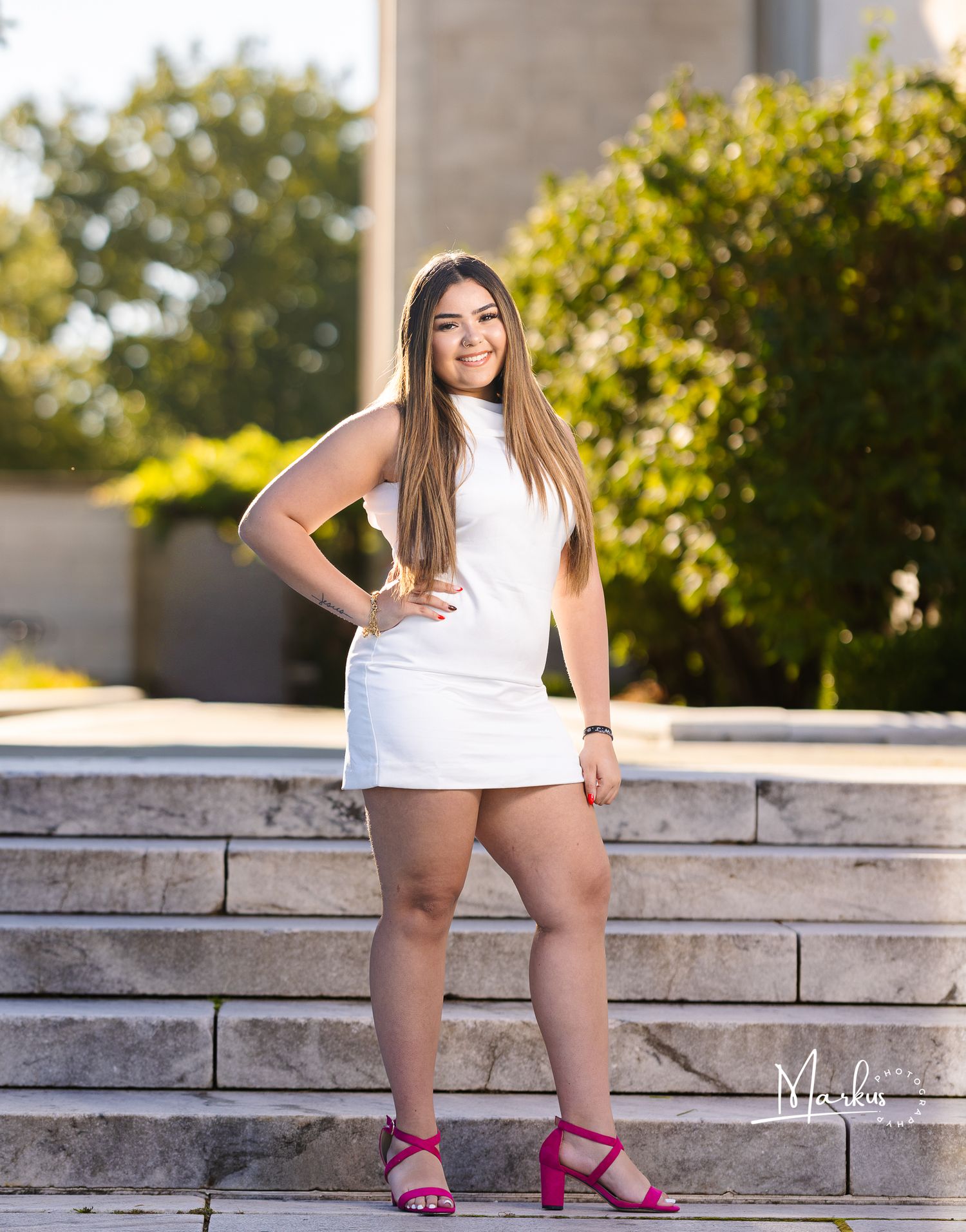 Young woman in white mini dress and pink heels posing confidently on stone steps outdoors.