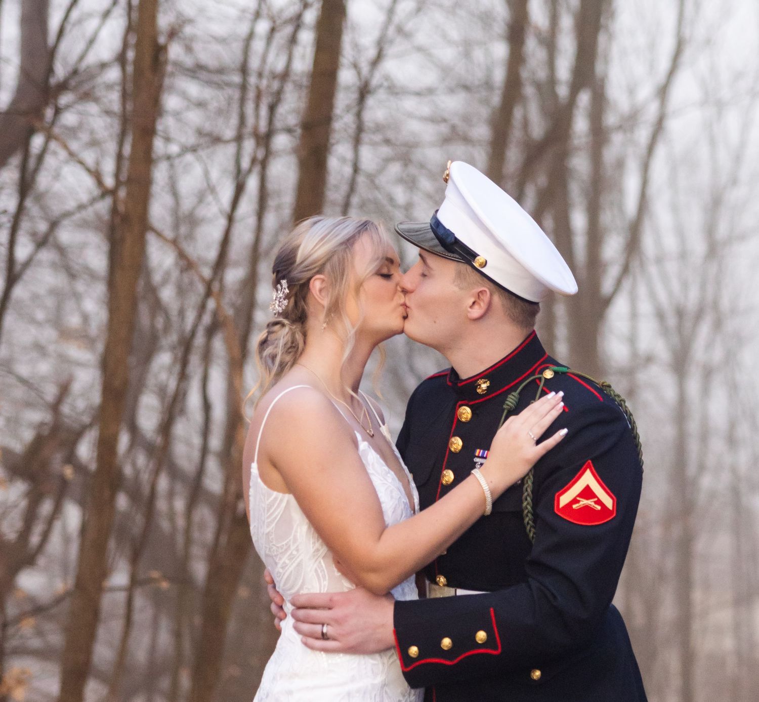 A couple in military dress uniform and wedding attire share a romantic kiss in a wooded outdoor setting.