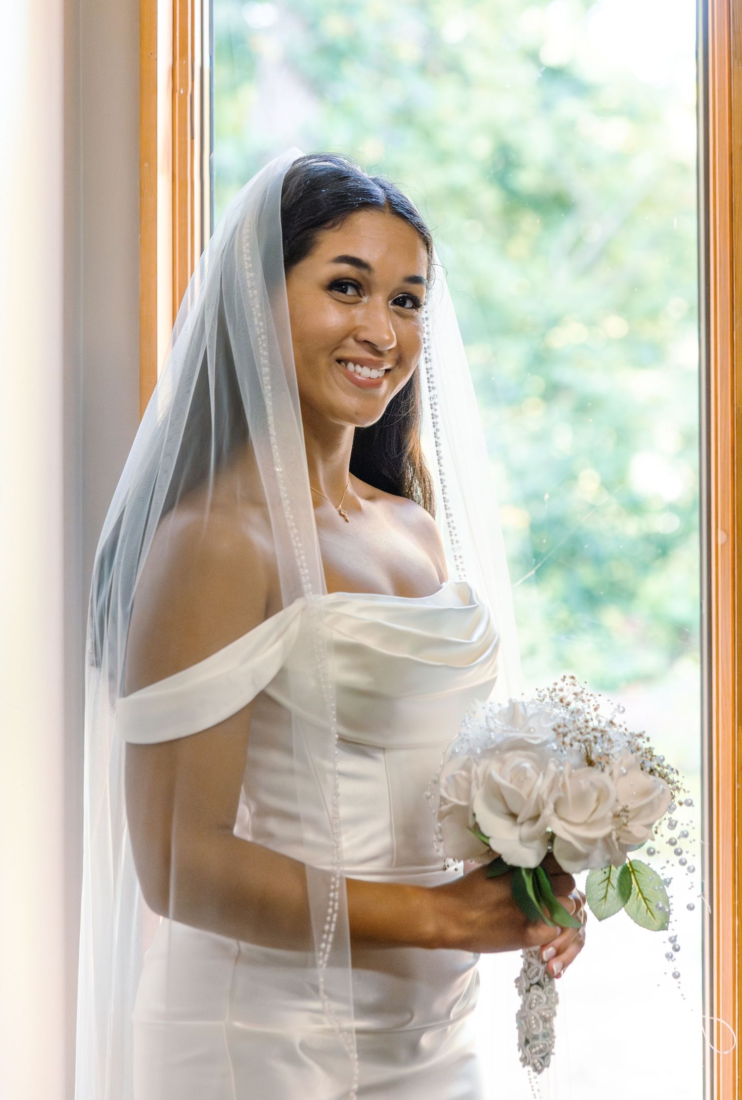 A bride in a white wedding dress and veil holds a bouquet of white roses while standing near sunlit windows.