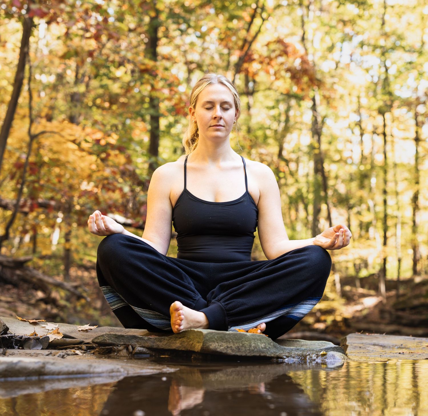 Person in black athletic wear sits in lotus position for meditation by a tranquil stream in autumn forest setting.
