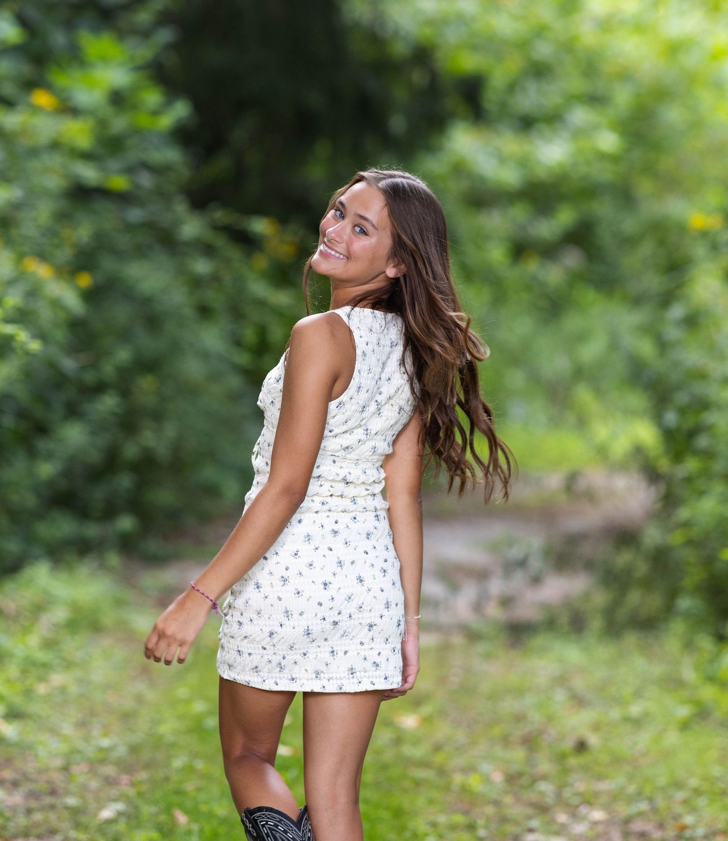 A person in a white polka dot dress dances playfully outdoors in a green summer garden setting.