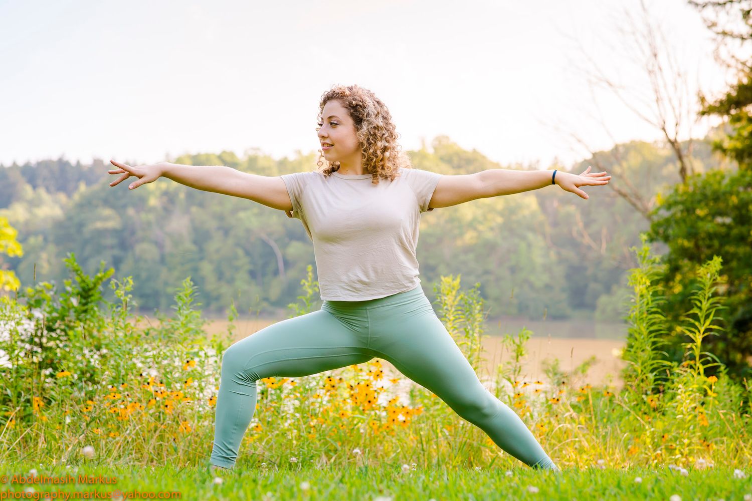 A woman in teal leggings practices yoga warrior pose outdoors in a sunny meadow with wildflowers.