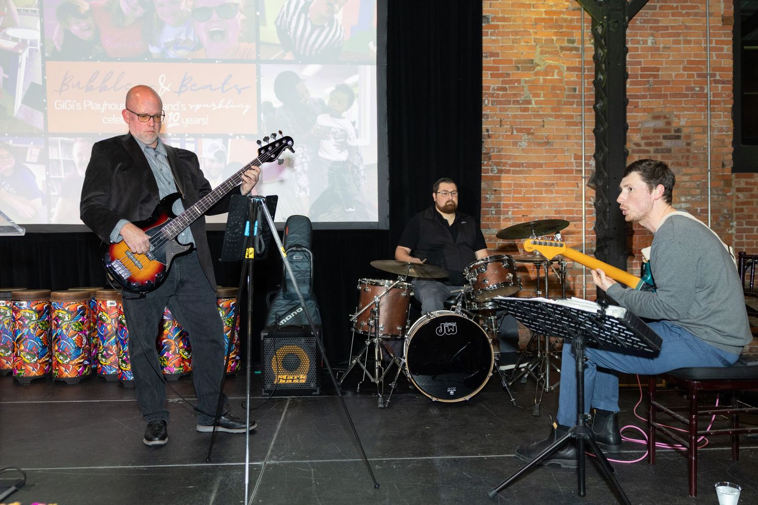A band performs on stage with a guitarist, drummer and keyboard player against a brick wall backdrop.