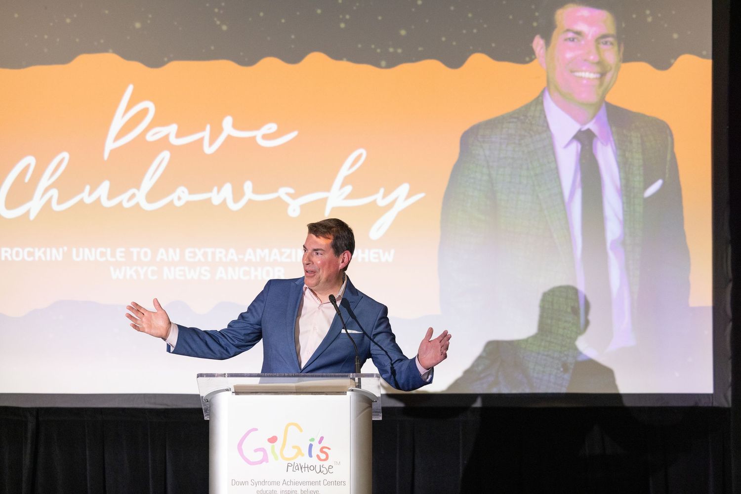 A presenter speaks at a podium during a conference with their image projected on screen behind them.