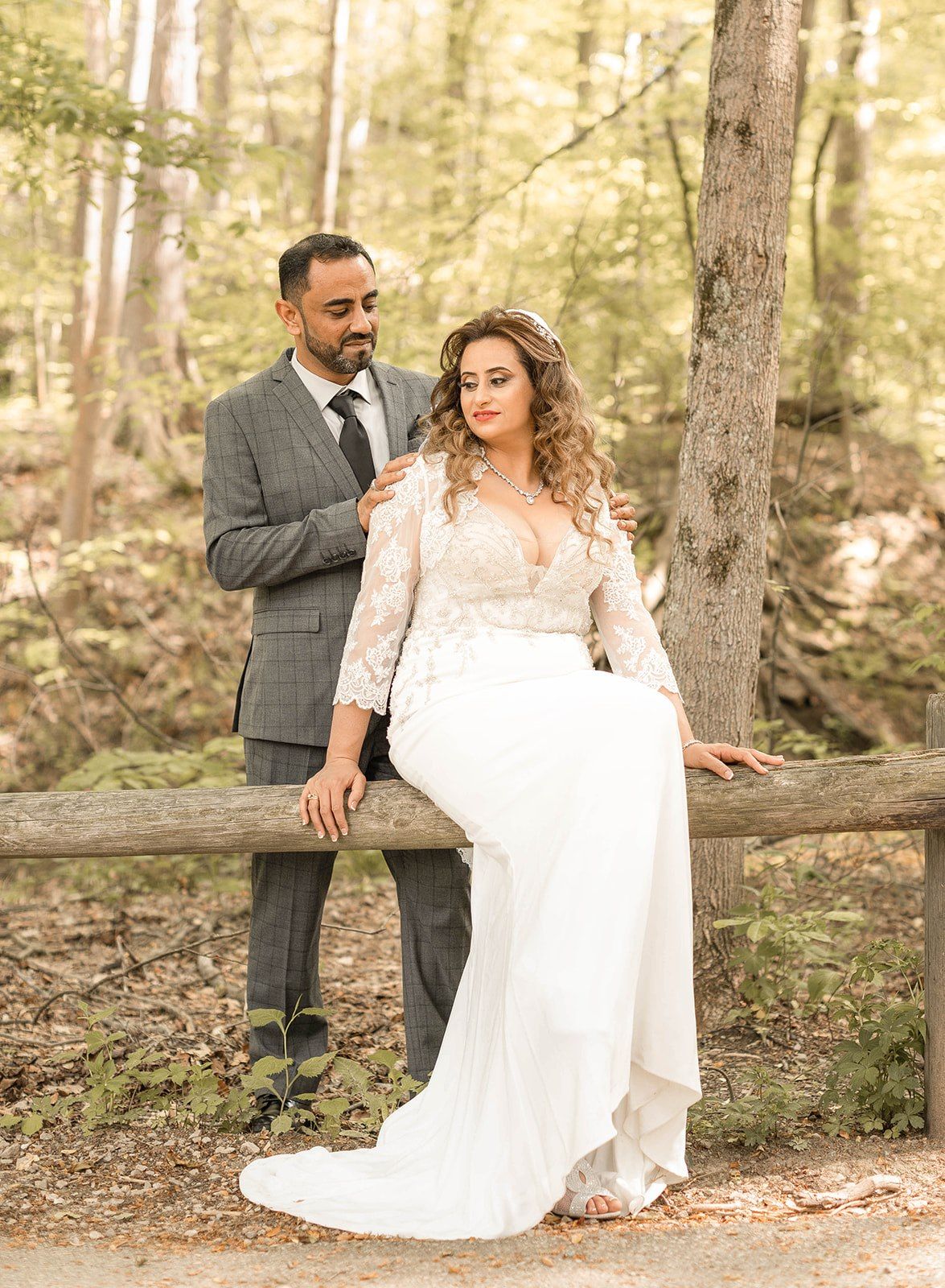 A romantic wedding couple poses together on a rustic wooden fence in a sunlit forest setting.