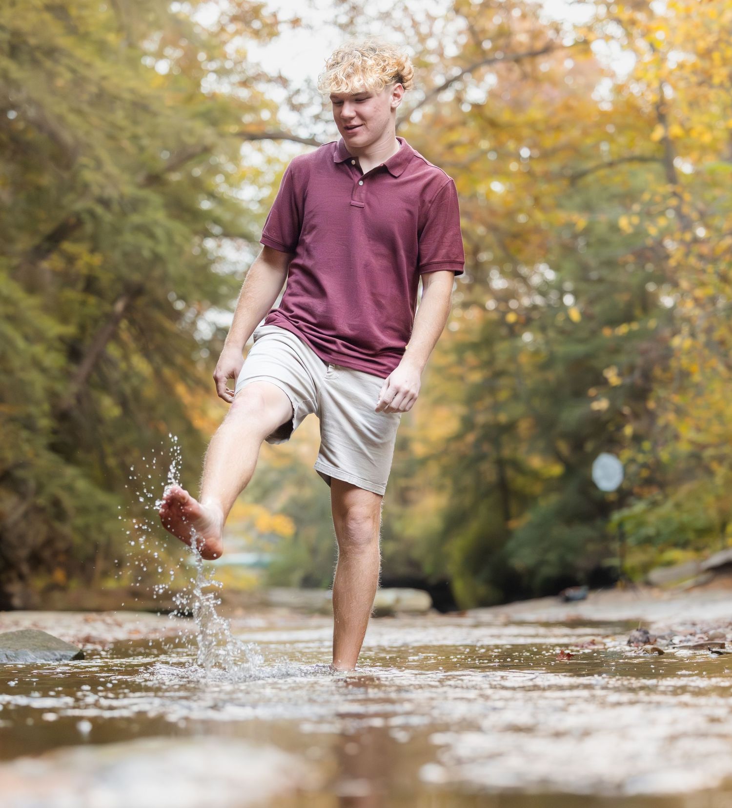 Person in burgundy polo shirt splashes through shallow creek water on autumn day with golden foliage in background.