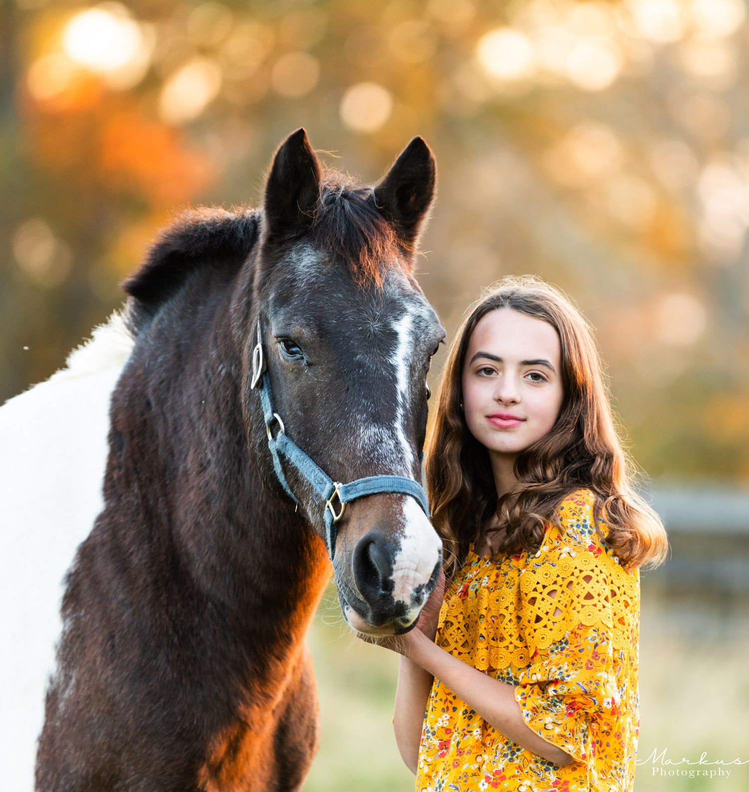 A black horse and person in a yellow floral dress stand together in a sunlit field during golden hour.
