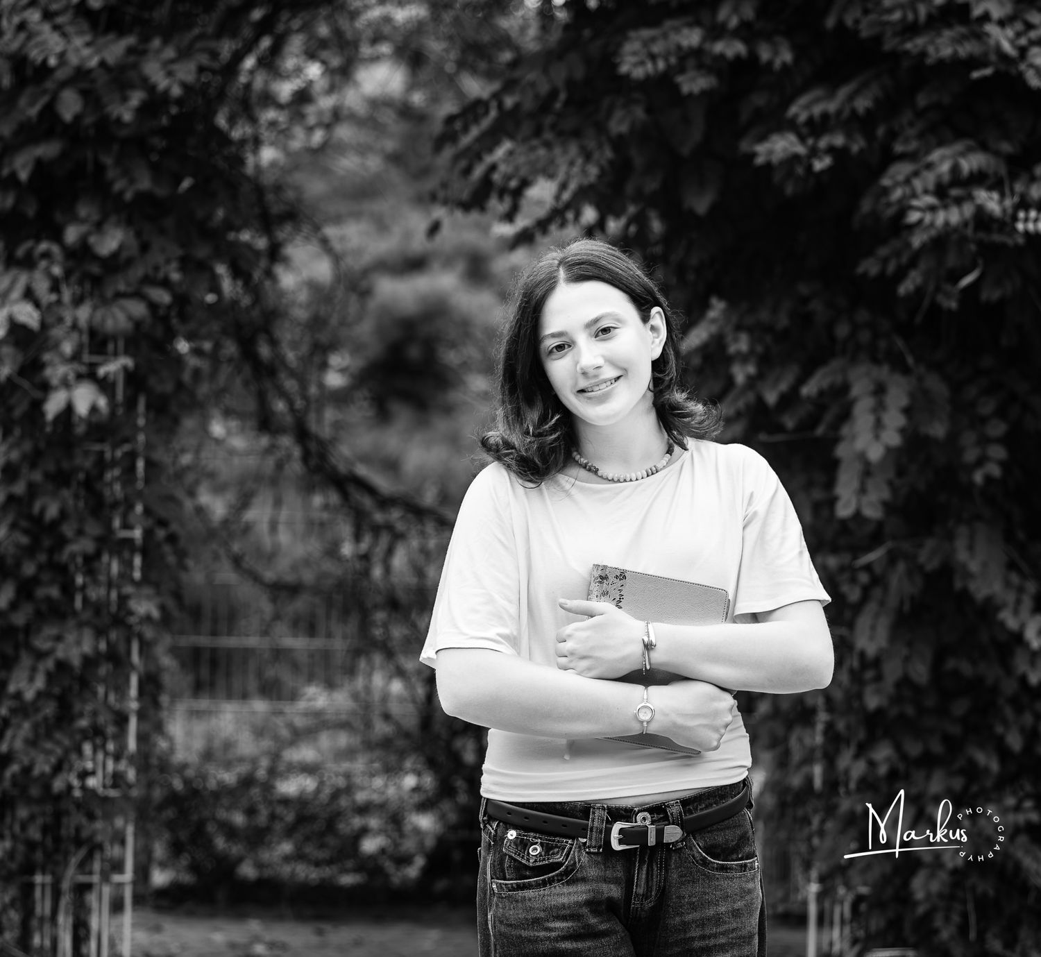 Black and white portrait of a young woman smiling in a garden, holding a book, wearing jeans and a white t-shirt.