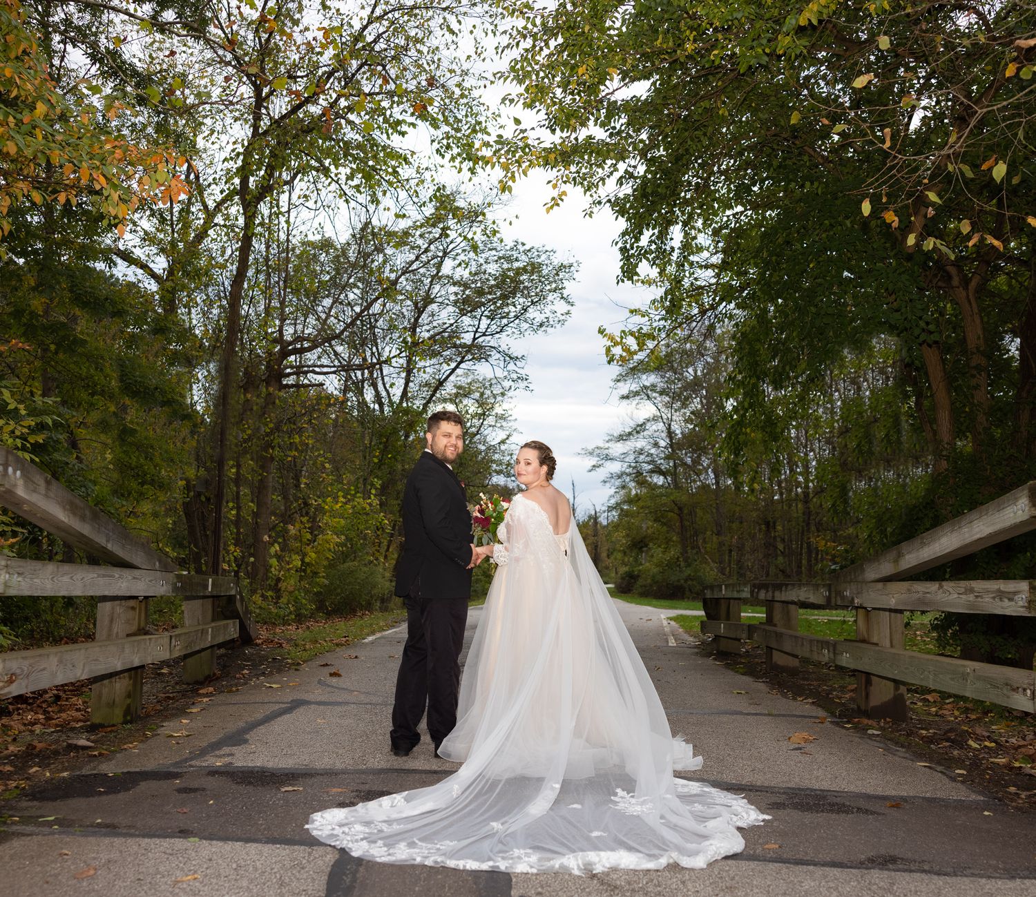 A wedding couple walks along a scenic tree-lined path in autumn with their flowing gown and formal attire.