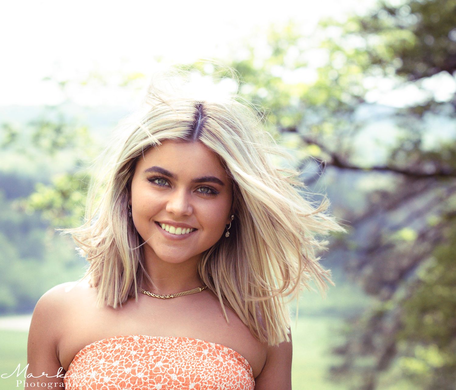 A person wearing a coral-colored top poses outdoors with natural greenery in the background.