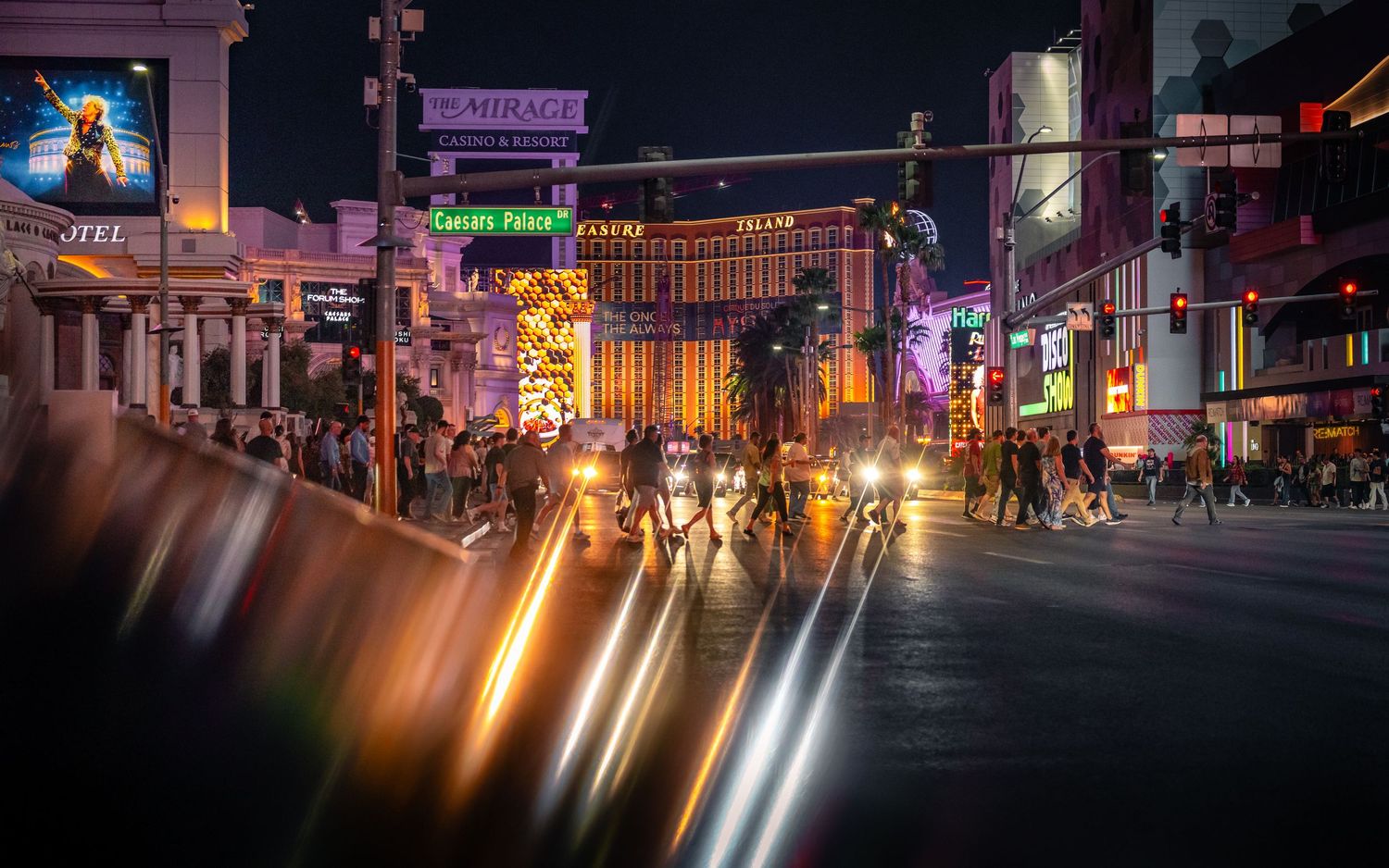 Blurred headlights and neon lights stretching across a Las Vegas street as pedestrians cross.