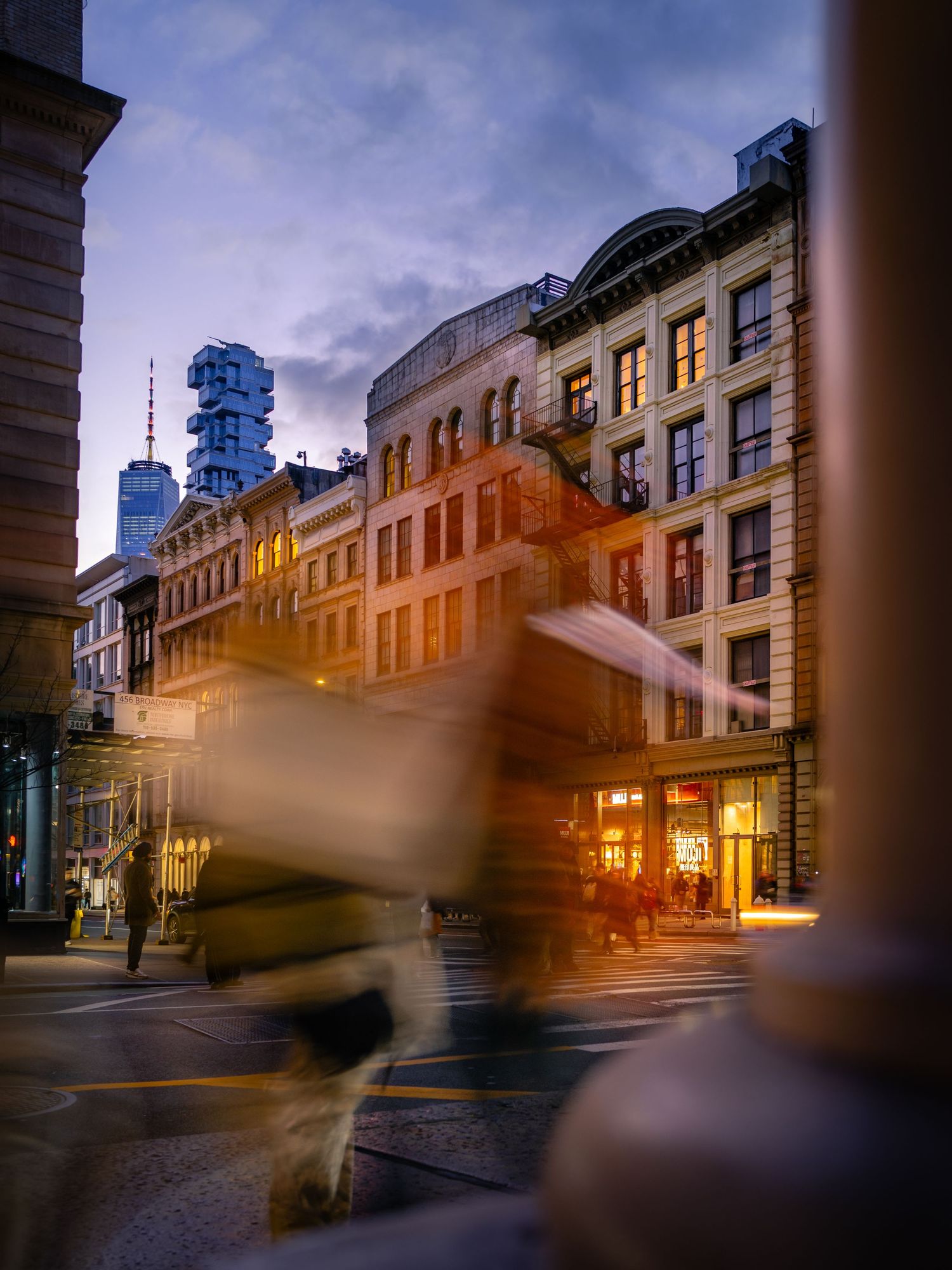 Long-exposure blur of people walking past storefronts in SoHo at dusk.