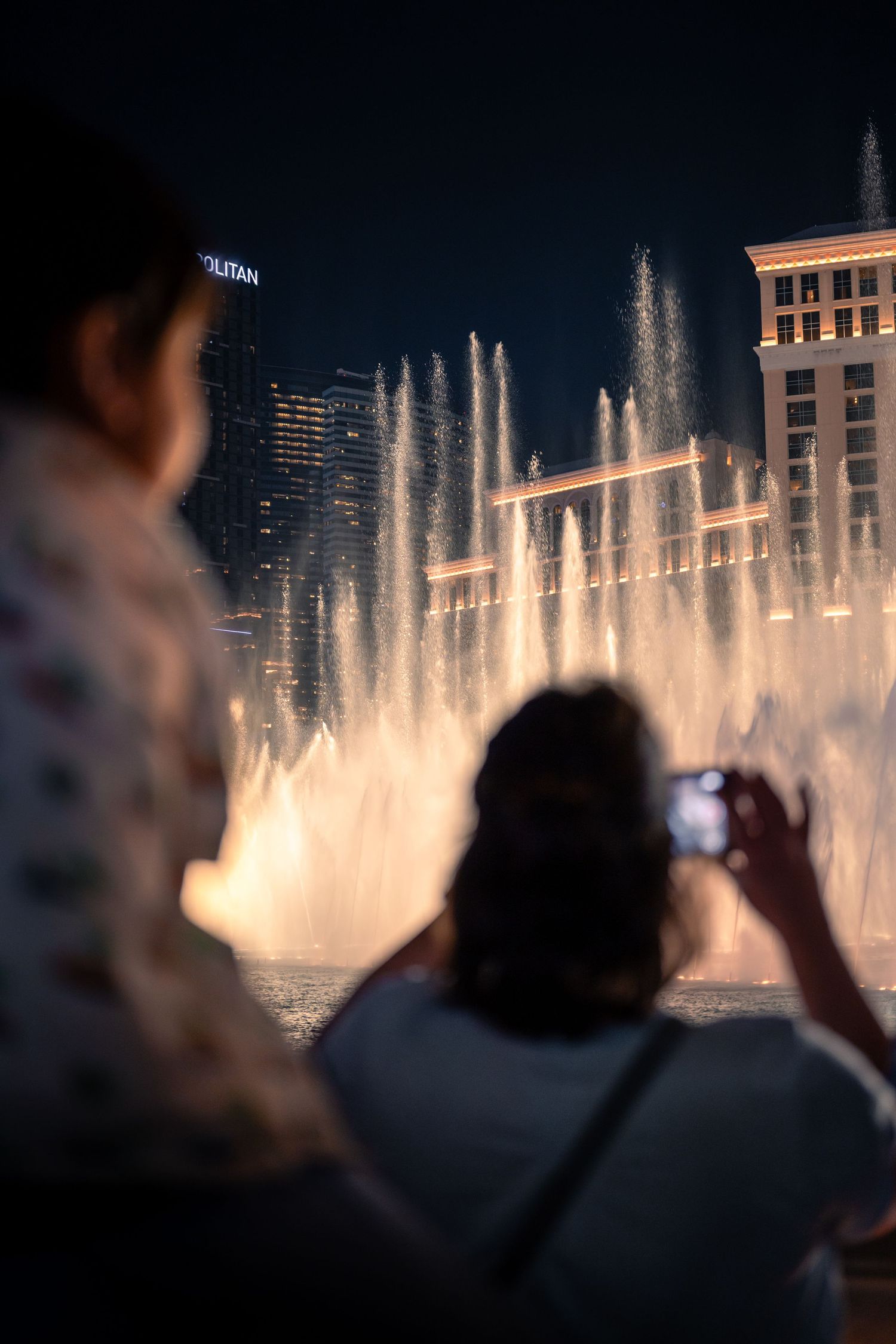 A child sitting on someone’s shoulders as a woman photographs the Bellagio fountains at night.