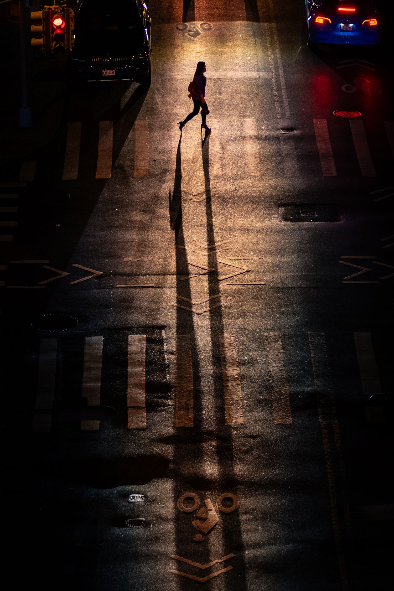 A woman crossing a city street at night, illuminated by streetlights and casting long shadows.