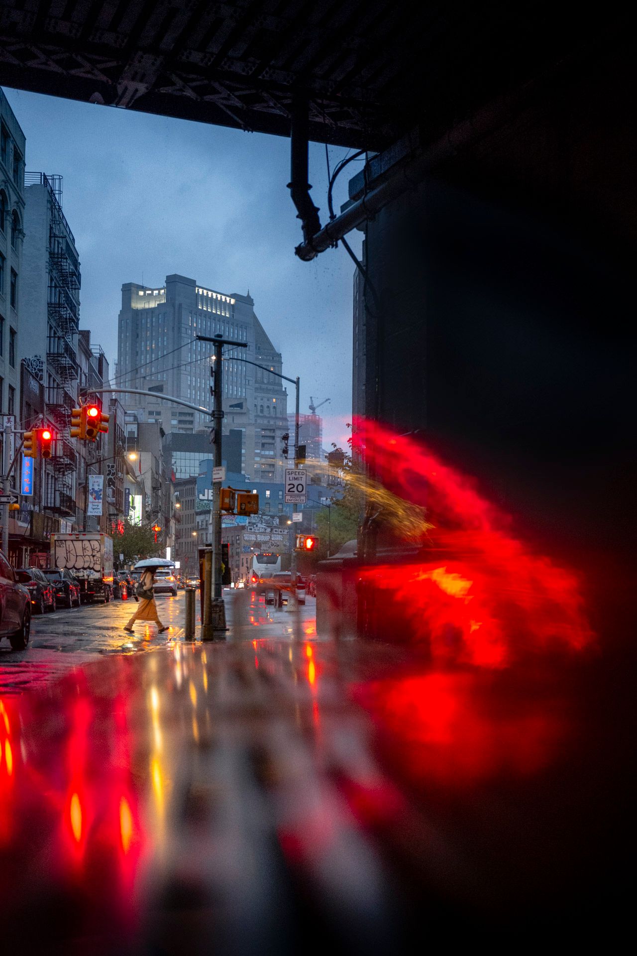 Pedestrian with an umbrella crossing a rain-soaked Chinatown street beneath a bridge with red light reflections