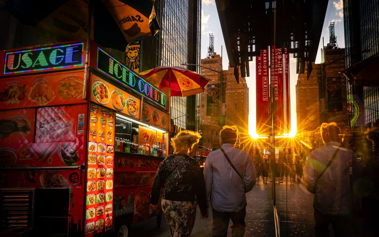 Pedestrians walking past a hot dog stand in Times Square as sunlight cuts through the street.