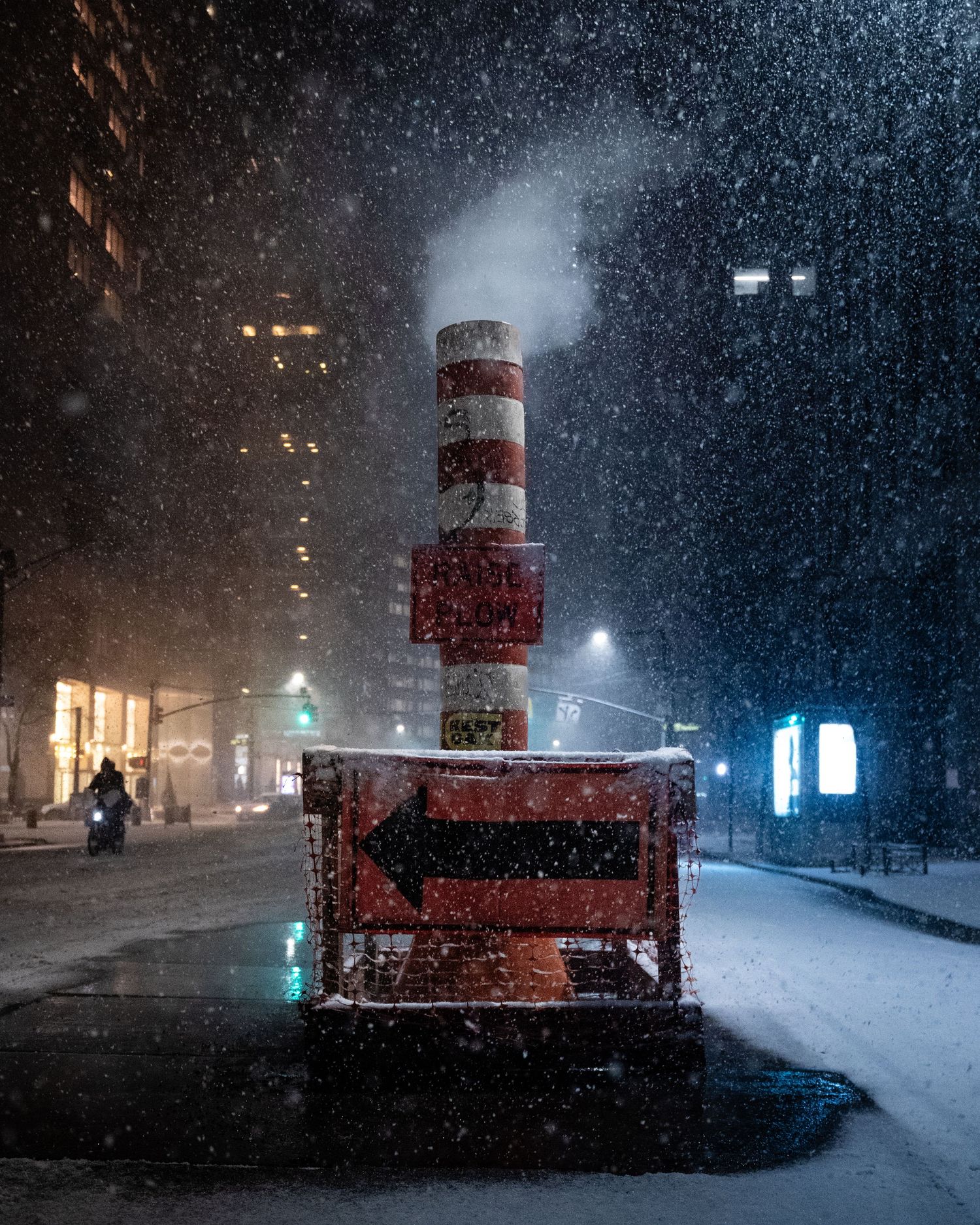Steam rising from a street vent, lit by yellow and blue lights on a snowy New York City night.