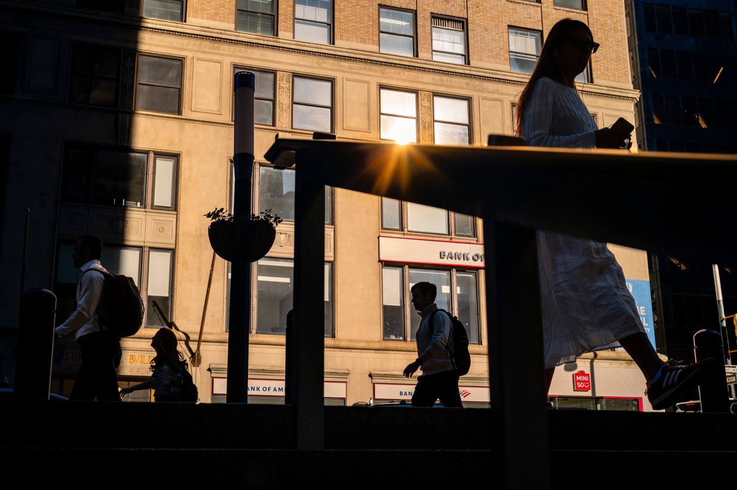 Pedestrian silhouettes framed by railings and shadows in warm evening light on a New York City street