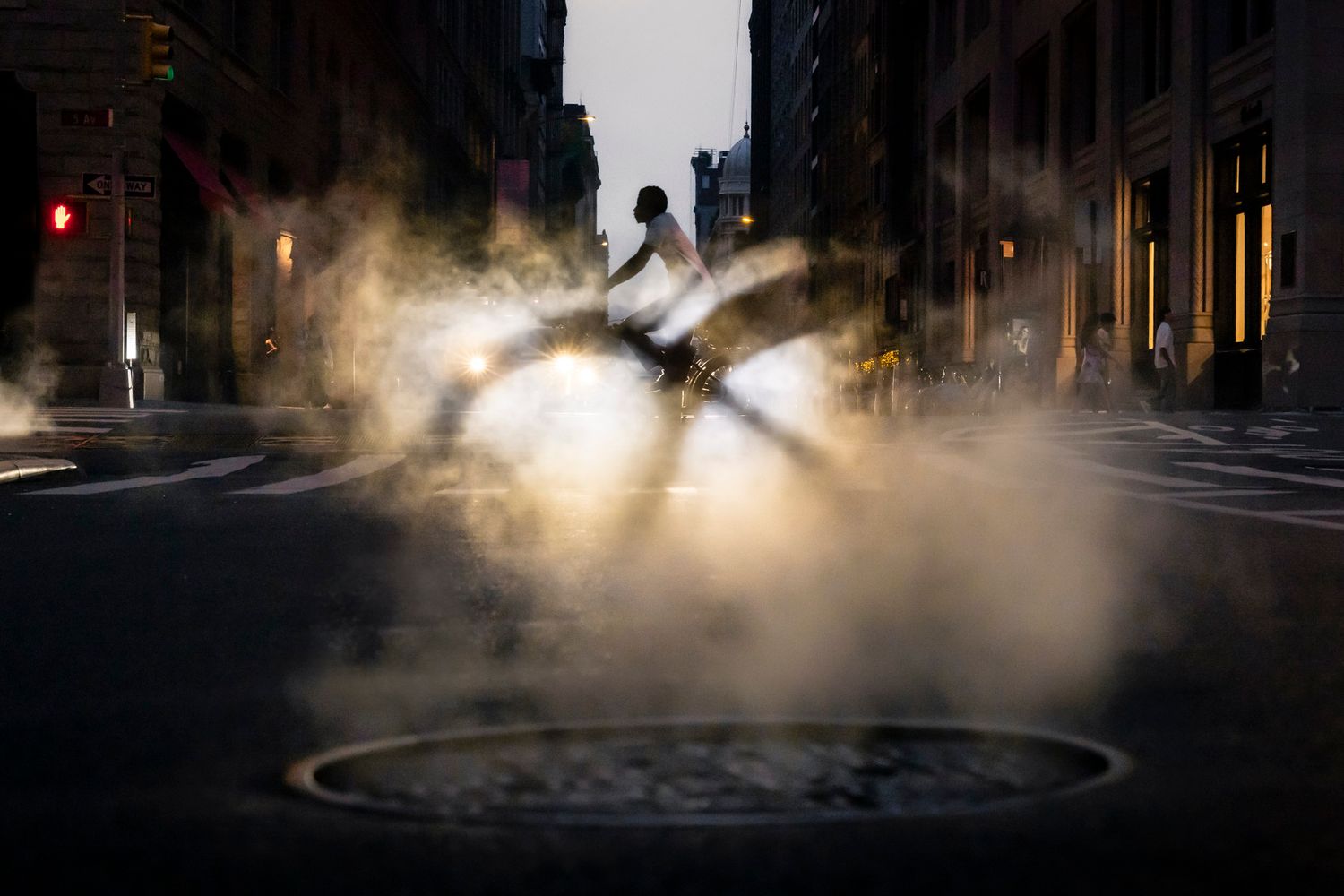 Cyclist silhouetted against headlights while riding through rising steam on a New York City street