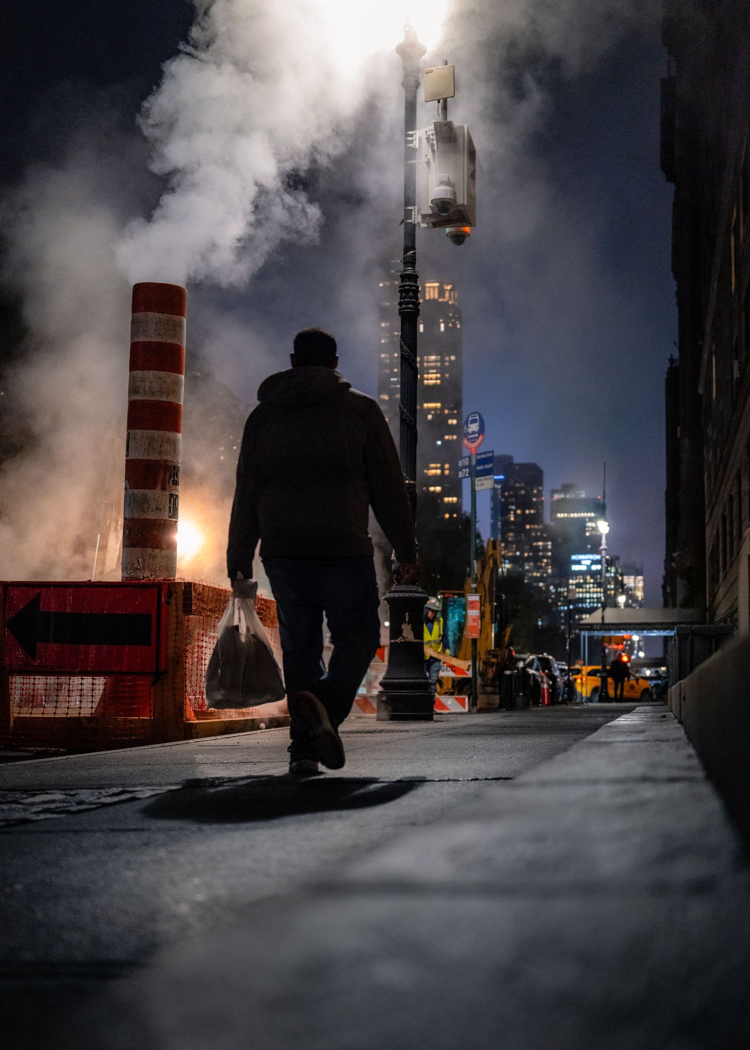 Silhouetted figure moving through street steam on a New York City sidewalk at night.