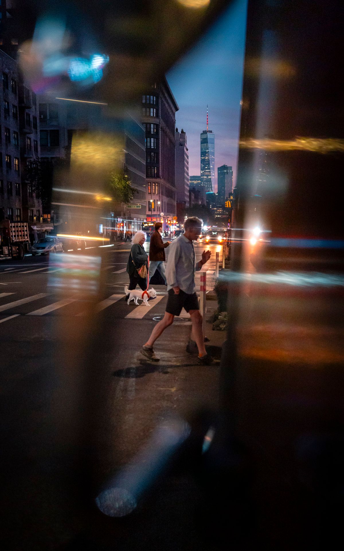 Pedestrians crossing the street through colorful reflections at dusk with One World Trade Center in the background