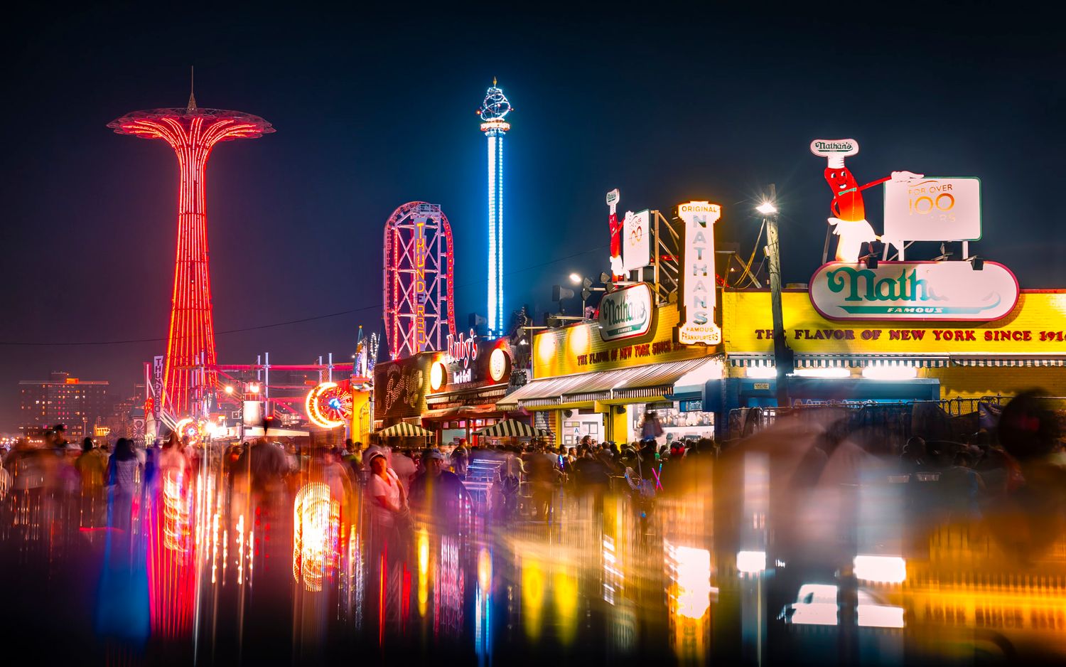 Coney Island boardwalk and amusement rides glowing at night with colorful reflections and motion blur