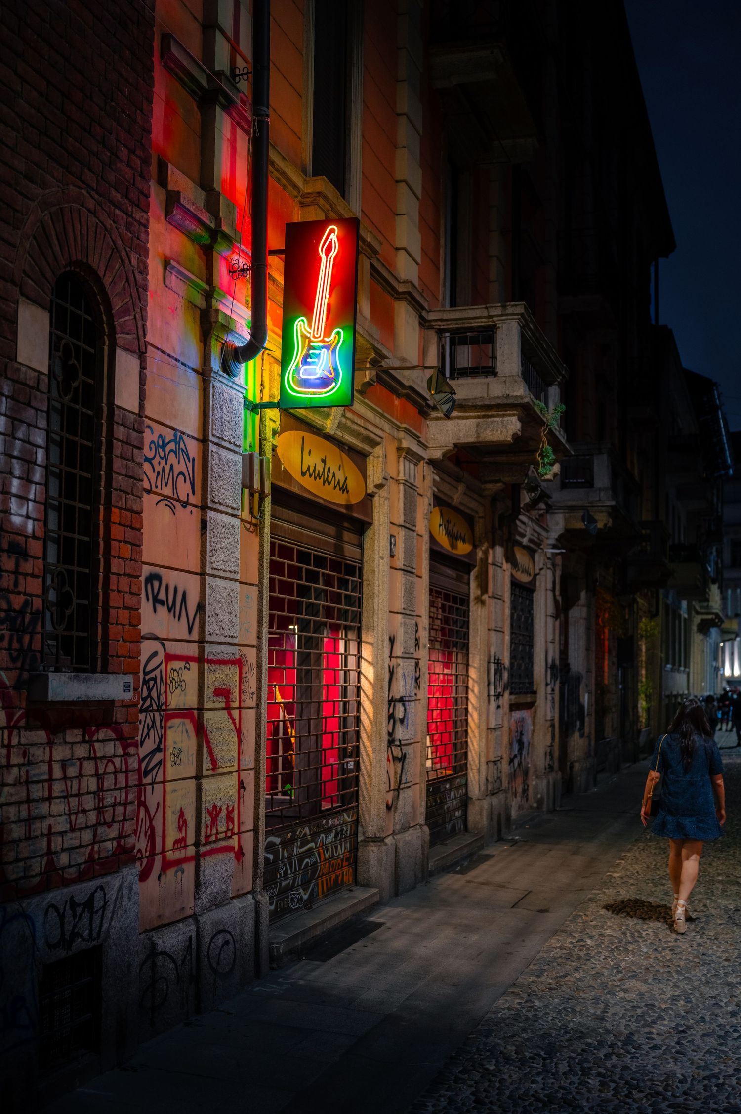 Neon lights glowing above a woman walking alone through a Milan street at night.
