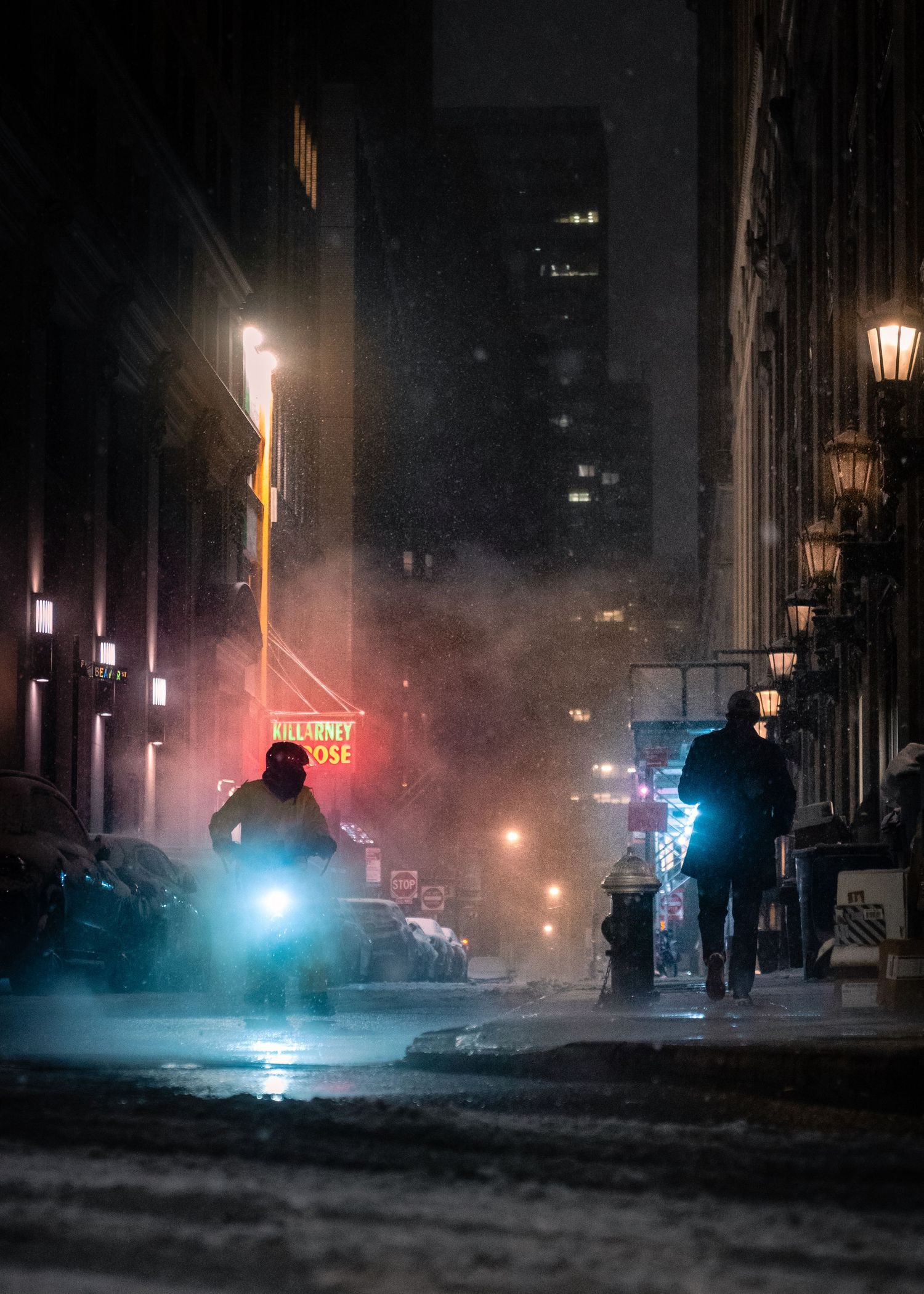 A delivery cyclist riding through falling snow on a dark New York City street at night.