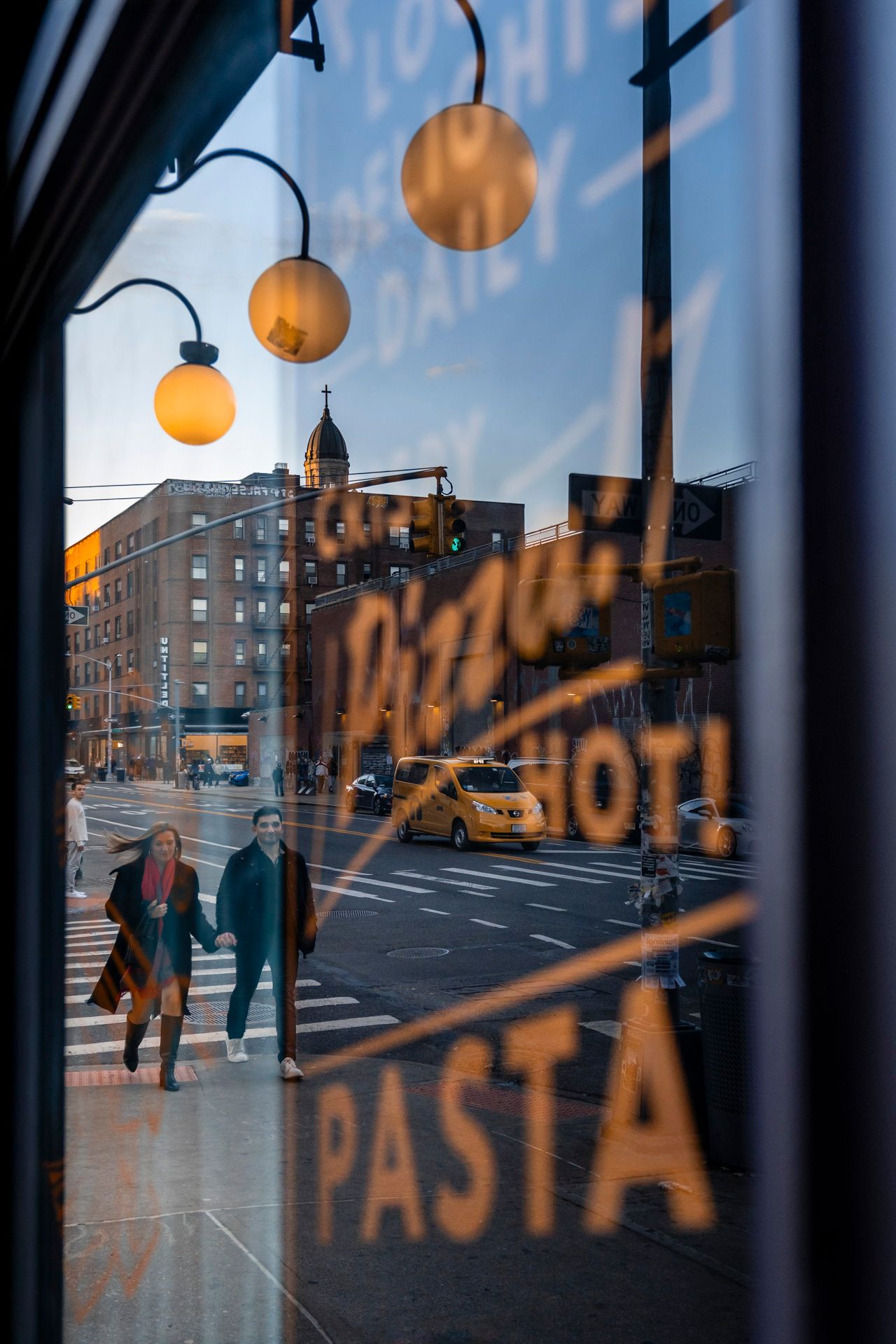Pizza and pasta shop window reflection framing couple holding hands and a yellow taxi on a New York City street.