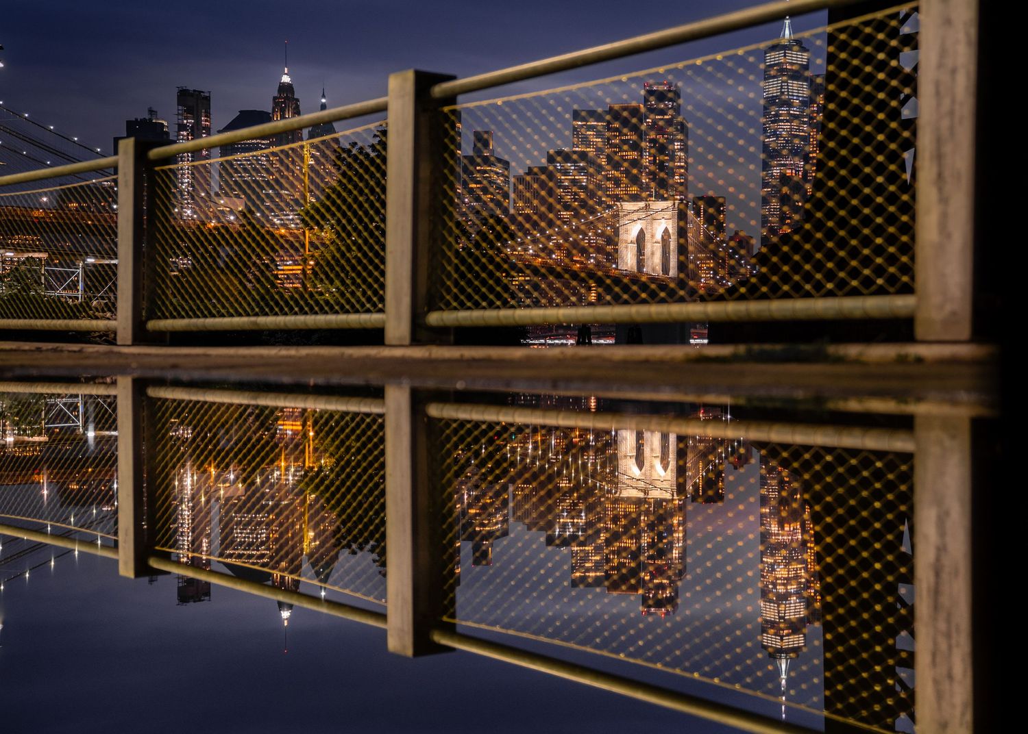 Reflection of the Brooklyn Bridge and Manhattan skyline mirrored in still water at night.